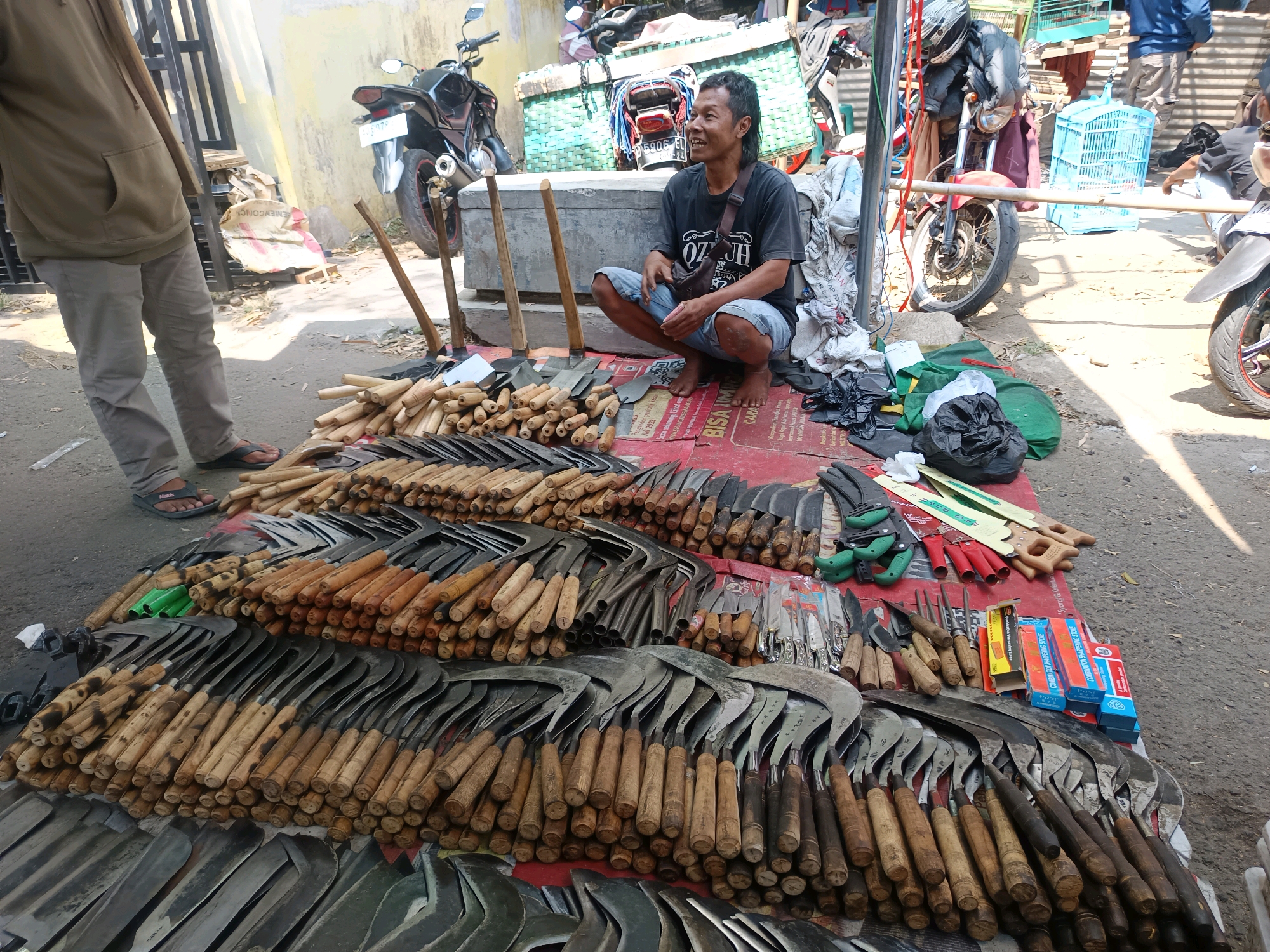 The image shows an outdoor market stall in Geneng, Indonesia, where a man is selling agricultural tools. He is kneeling on a red mat, surrounded by an array of machetes, sickles, knives, and other metal implements with wooden handles. Some of the tools are laid out in neat rows, while others are stacked in bundles. A person in a brown hoodie stands in the background to the left, partially obscured by a vertical pole. In the background, motorcycles are parked, and various other market items, including crates and bags, are visible. The ground is paved with asphalt. The scene appears to be a typical street market in a rural or semi-urban setting.