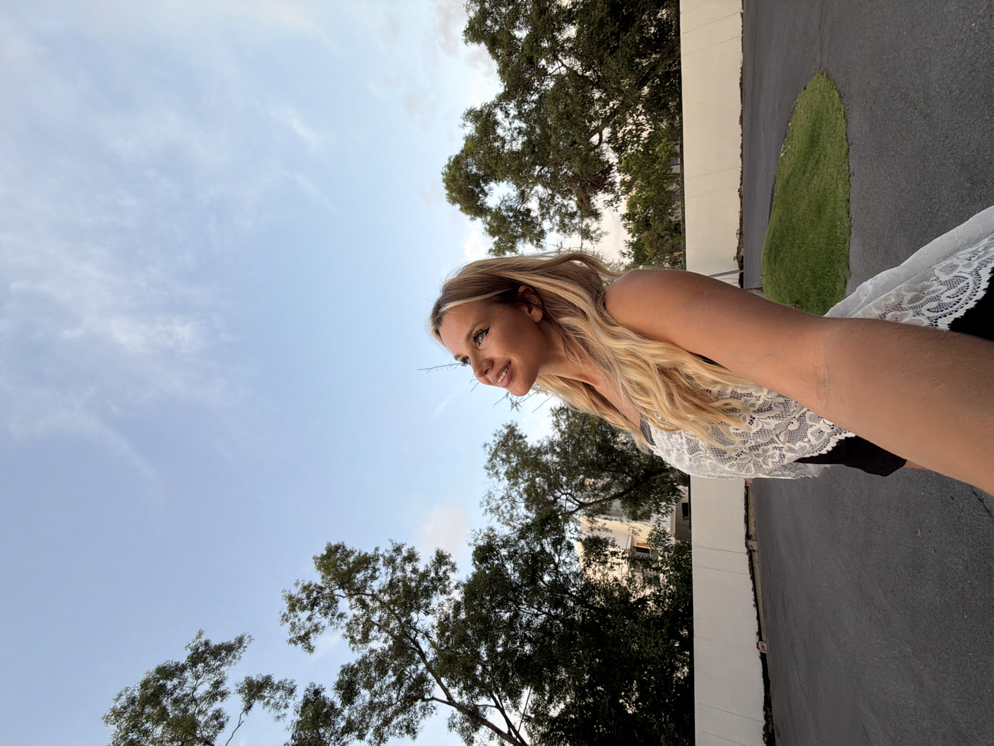 A blonde woman is seen from the chest up, looking towards the sky and smiling. She is wearing a white lace top over a black garment. The background consists of trees, a white wall, and a paved area that appears to be a road or parking lot. The sky is blue with some scattered clouds. The location is identified as Thep Krasatti, Thailand.