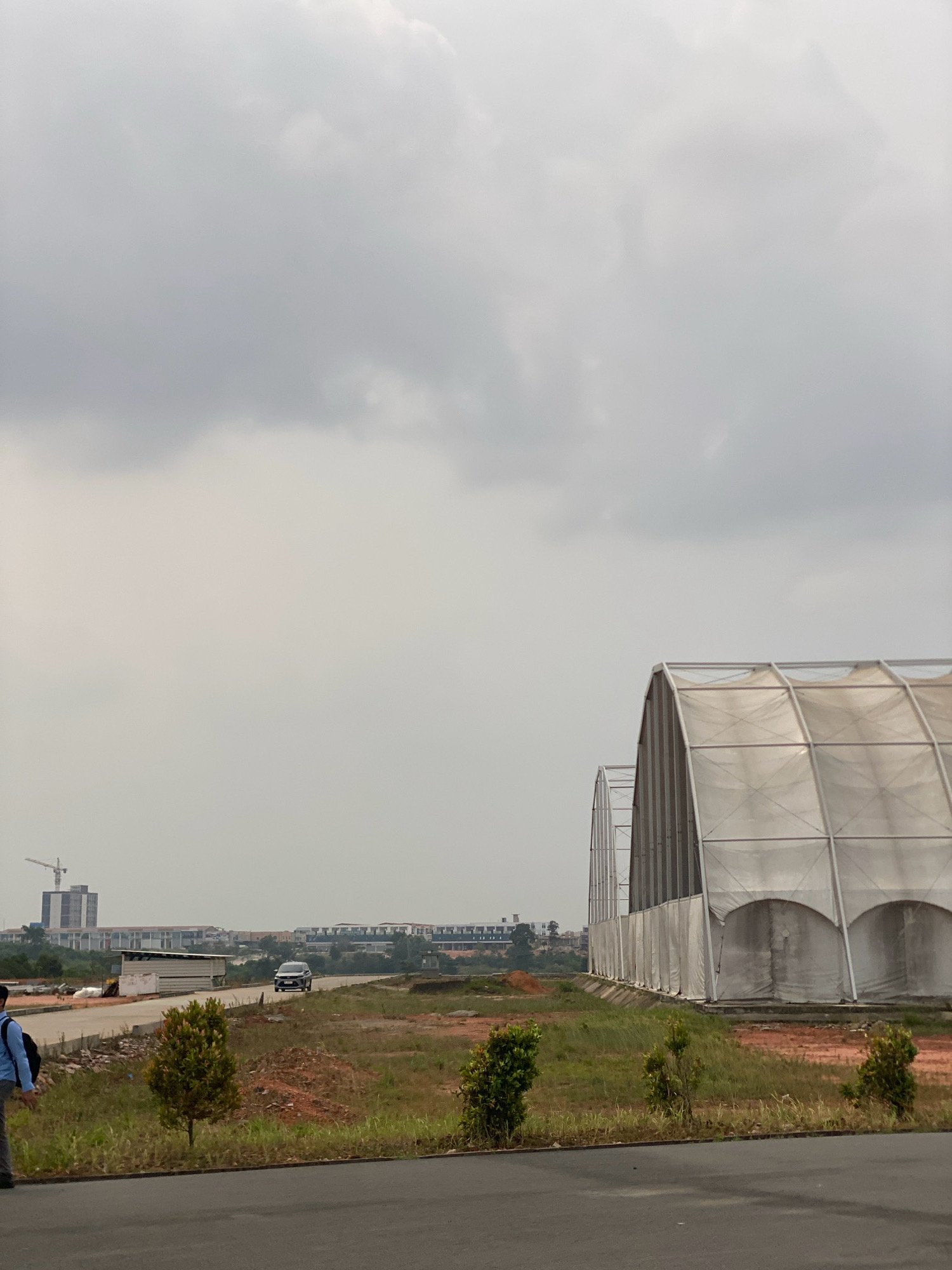 The image depicts an outdoor scene, likely in Batam, Indonesia, characterized by a cloudy sky and an overcast atmosphere. In the foreground, a person dressed in blue and grey attire with a backpack is walking on an asphalt surface. Beyond the asphalt, a patch of dry, reddish soil is visible, interspersed with sparse green grass and a few small trees. A paved road runs through the middle ground, where a dark-colored car is driving. In the background, a long, white, curved structure, possibly a temporary building or tent, dominates the right side of the frame. Further in the distance, a construction crane is visible near a cluster of tall buildings, suggesting ongoing development in the area. The overall environment appears to be a developing or industrial zone.