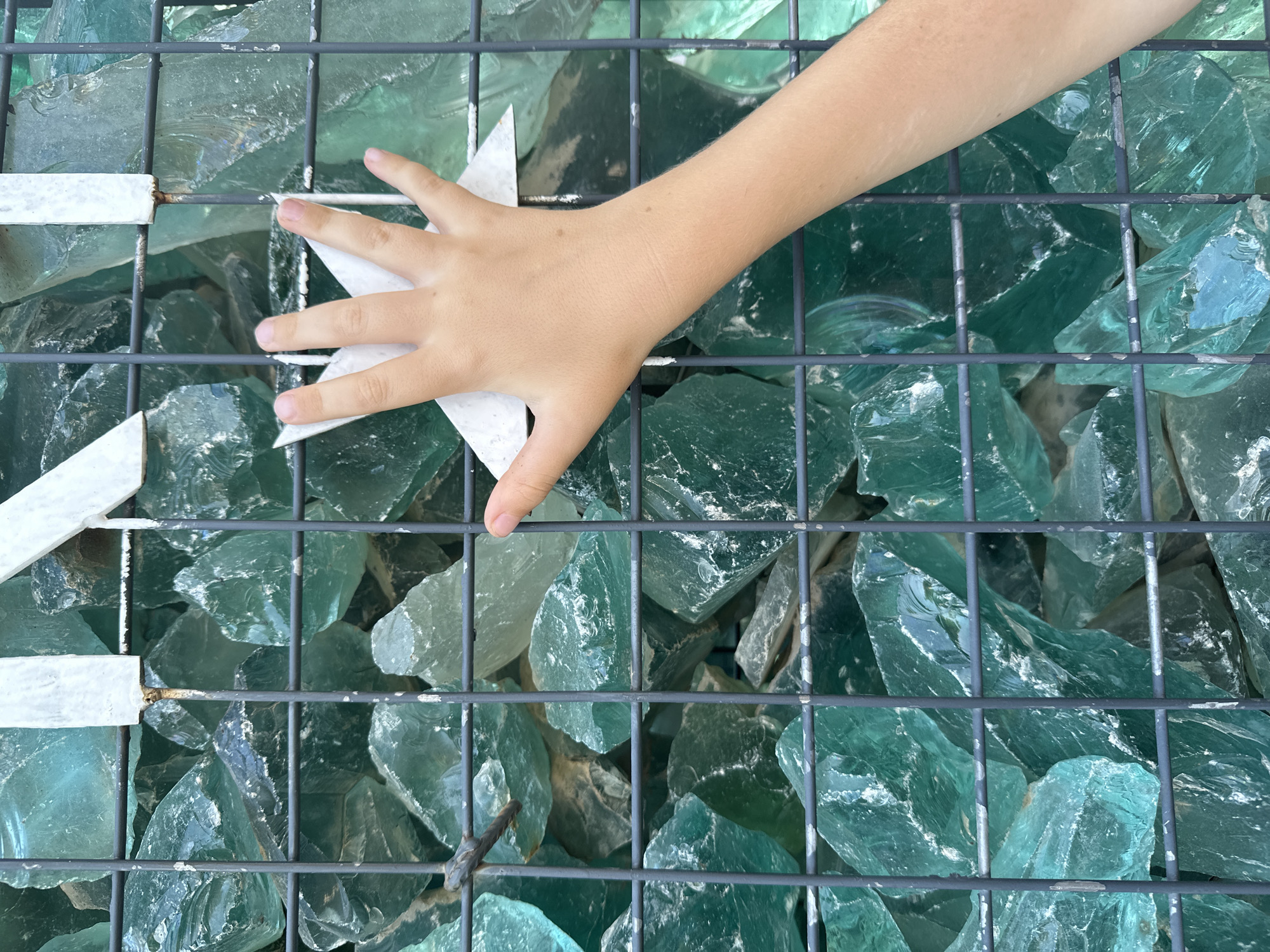A child's left hand, fingers spread, is pressed against a white, star-shaped object, which is attached to a metal grate. Behind the grate, a dense collection of large, irregularly shaped pieces of translucent, teal-colored glass are visible. The glass appears to be piled high, creating a textured and layered background. The scene is lit by natural light, casting subtle shadows on the glass and hand. This arrangement is located in Fethiye, Turkey.