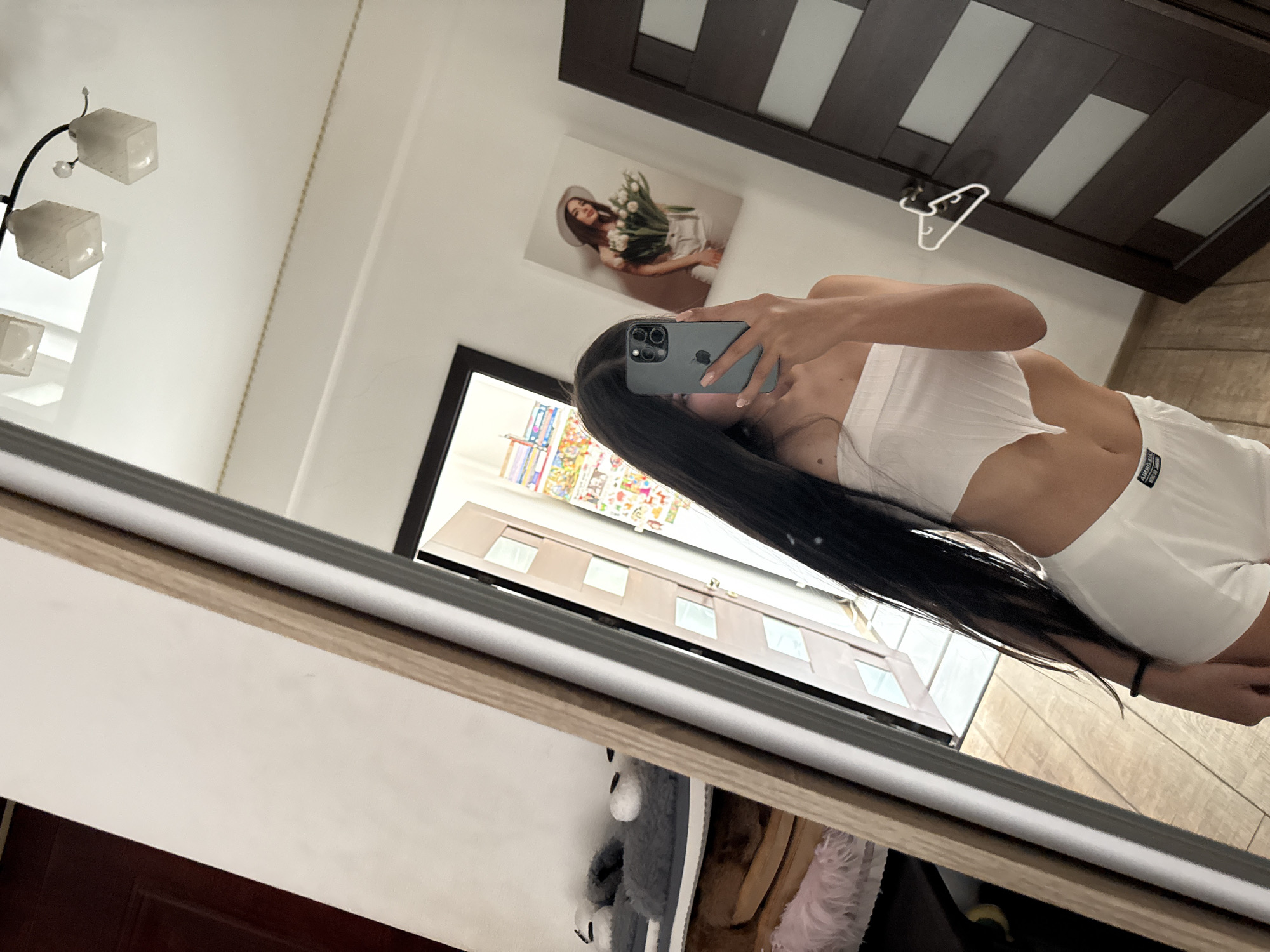 The image shows a young woman with long, dark hair taking a selfie in a bedroom. She is wearing a white strapless top and white shorts. Her phone is held up to take the picture, and her face is partially obscured by it. In the background, a mirror reflects the room, revealing a doorway, a dark wooden door, and a framed picture on the wall. The room appears to be a bedroom with wooden flooring and light-colored walls. There are also some slippers and a fluffy item on a shelf to the left of the mirror, which are partially visible. There is a light fixture with multiple glass shades on the ceiling. The scene does not provide any specific indicators to determine its location within Chișinău, Moldova.