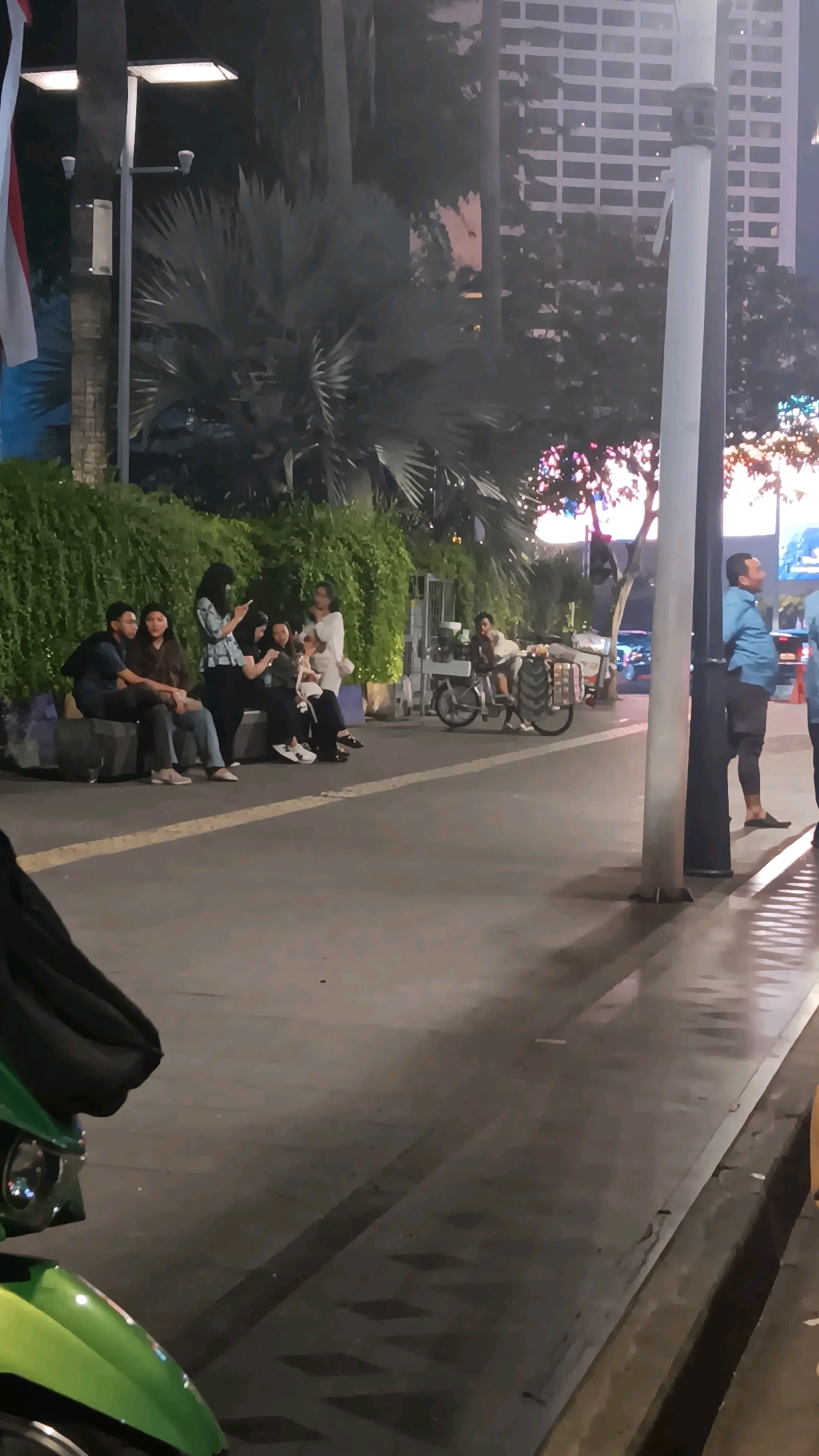 The image captures an outdoor scene at night in Central Jakarta, Indonesia, likely a public space like a promenade or sidewalk area near a building. In the foreground, a green scooter is partially visible. Further back, a group of six young adults, appearing to be four women and two men, are seated on a low wall or platform. They are casually dressed and engaged in conversation or looking at their phones. To the right of this group, a bicycle with a cart or basket attached is parked. A man in a blue shirt and dark shorts stands to the right, looking away from the camera. The background shows lush green foliage and palm trees, illuminated by artificial lights. Tall buildings, characteristic of a city environment, are visible in the distance, their windows lit up. The overall ambiance suggests a relaxed evening gathering in an urban setting.
