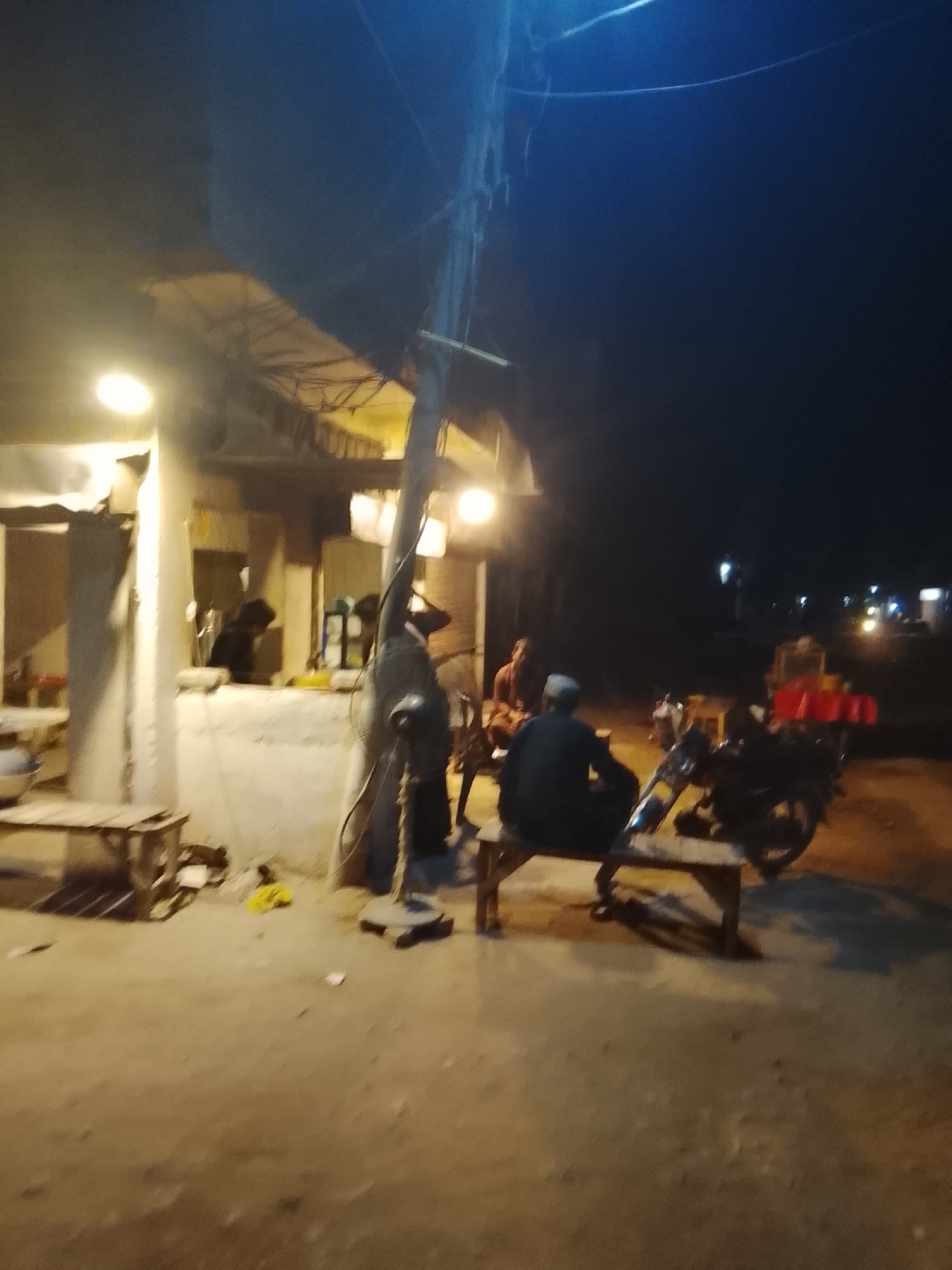 This is a blurry, nighttime photograph depicting an outdoor gathering scene, likely at a small eatery or tea stall. The sky is dark, indicating it is late evening or night, with the primary illumination coming from several bright, warm-toned lights within and around a simple, open-fronted structure on the left.
Several people are present. In the mid-ground on the right, a person wearing a dark top and a light cap is seated on a wooden bench, facing away from the camera. Adjacent to this individual, a motorcycle is parked. To the left, another person is visible, seated on a similar wooden bench, facing towards the center. Inside the illuminated stall on the left, two more figures are dimly visible, one appearing to be attending to a counter. The individuals seem to be socializing or relaxing in this communal space.
A large utility pole stands prominently near the center of the frame, with electrical wires extending upwards. A standing fan is visible at the base of this pole. The ground is unpaved, appearing to be dirt or compacted earth, with some scattered items. In the distance on the right, blurry lights suggest other structures or vehicles. No discernible text is visible due to the blurriness and low light conditions. The overall atmosphere appears casual and laid-back.