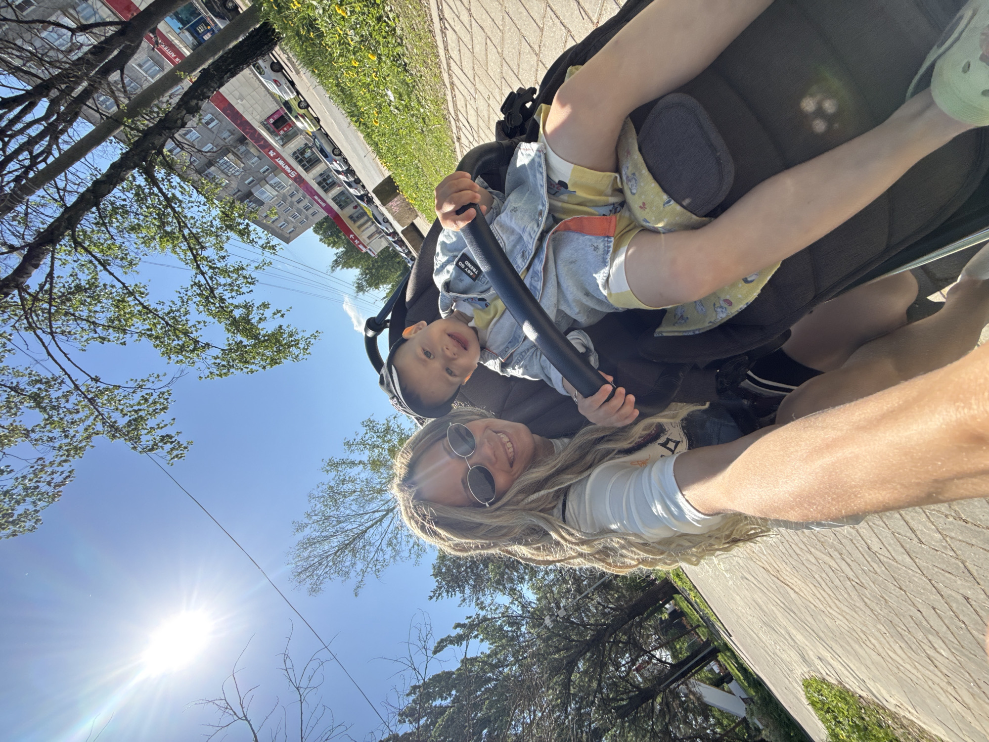 A woman in sunglasses is holding a camera up, taking a selfie, with a baby in a stroller. The baby is wearing a light blue denim jacket and yellow shorts, holding onto the stroller’s bar. They are outside on a sunny day, with trees and buildings visible in the background. The location appears to be a city street.