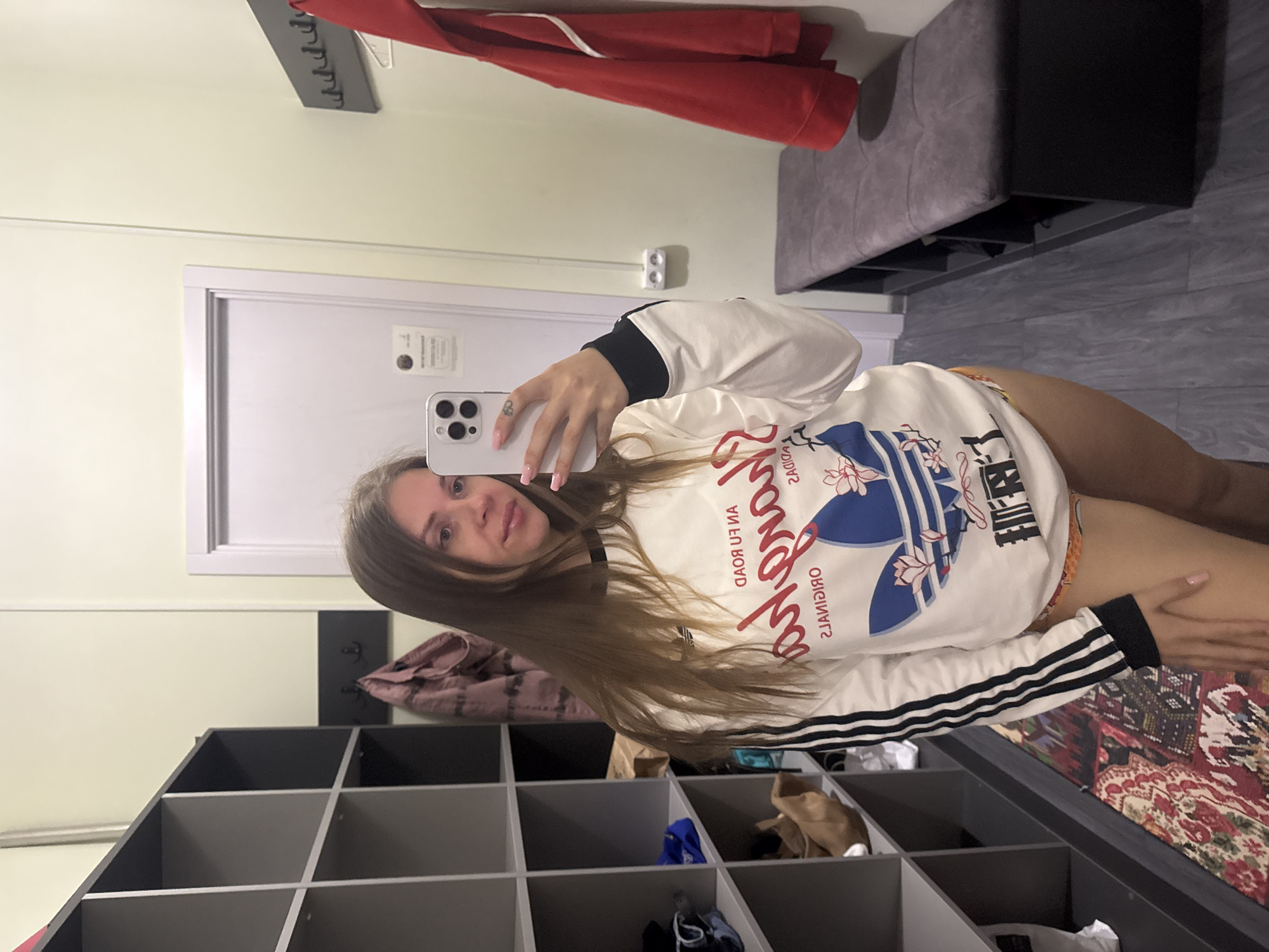 A woman with long blonde hair is taking a selfie in a dressing room. She is holding an iPhone with her right hand. She is wearing a white Adidas sweatshirt with black accents. The dressing room includes a gray shelf with various items inside, a door, a coat rack with a red jacket hanging on it, and a bench. The floor is tiled. The location is Batumi, Georgia.