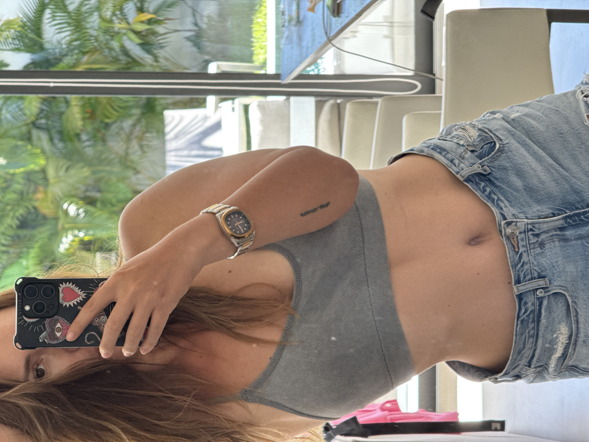 This is a vertical, mirrored selfie taken by a person holding a smartphone. The person is wearing a grey ribbed sports bra and distressed denim shorts. A watch with a gold and silver band is visible on their left wrist. There is a small tattoo on their inner forearm. The background appears to be an indoor setting with large windows reflecting greenery, suggesting a location with outdoor plants, possibly a patio or a room with a view of a garden. Chairs and a table are also reflected in the window, indicating a dining or living area. The lighting suggests daytime. The person's face is partially visible in the reflection on the phone screen, and their hair is long and brown. The smartphone case has a black background with a pattern that includes a red heart, an eye symbol, and a leopard print design.