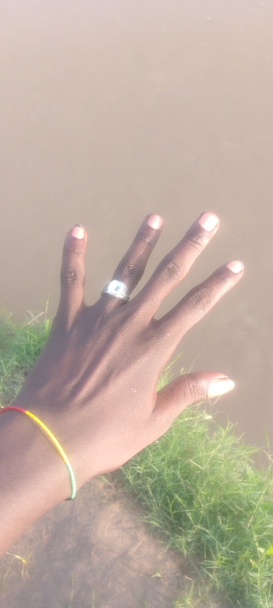 This media file is a close-up shot of a person's left hand, which is held up with fingers spread, against a blurred background of water and grass. The hand is tanned, and the nails are clean and lightly colored. On the ring finger, there is a silver ring with a square, clear stone. A bracelet, made of red, yellow, and green beads, is visible on the wrist. The background suggests an outdoor setting, possibly near a body of water, with green grass in the foreground and a smooth, muddy brown surface that appears to be water in the background. The lighting suggests it is daytime, with the sun casting highlights on the skin and the ring. No text is visible in the image. The focus is on the hand and its adornments, with a sense of calm or contemplation conveyed by the pose. The location context of Jalingo, Nigeria, adds a geographical specificity to the scene, suggesting a warm climate suitable for outdoor activities near water.