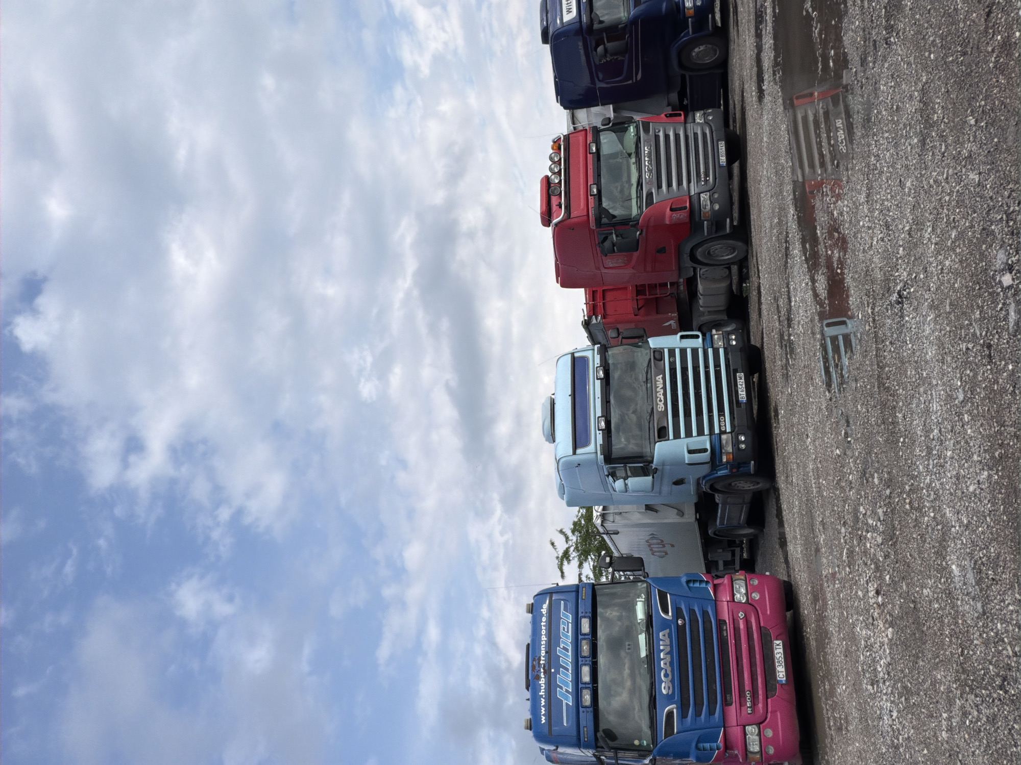 The image shows several Scania trucks parked on a gravel lot under a partly cloudy sky. The trucks are of varying colors, including blue, pink, light blue, red, and dark blue. The license plate on the pink/blue truck reads "CT 3853 TK" while the blue/black truck has "BT 6542 MB" and the red truck reads "BT 8559 KB". The words "www.huber-transporte.de" and "Huber" are visible on the side of one truck. The side of another truck also displays the word "argo". Based on license plates visible on the trucks the location is the city of Polski Trambesh, Bulgaria.