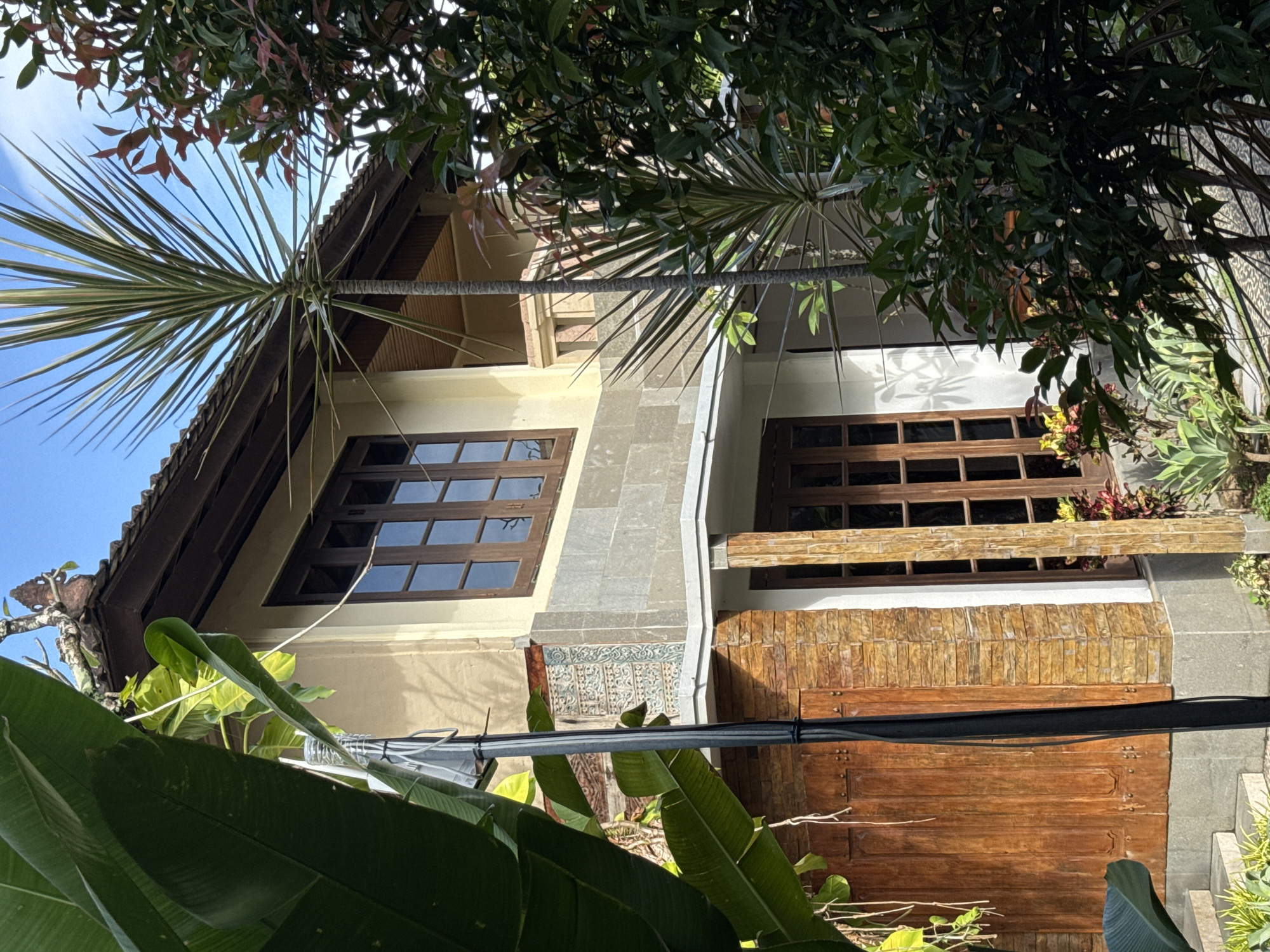 This is an outdoor, low-angle shot of a two-story building, likely a house or guesthouse, surrounded by lush tropical vegetation in Kuta, Indonesia. The image is captured on a clear, sunny day, with a bright blue sky visible in the upper left.
The building features a dark-tiled roof with overhanging eaves. The upper story has a light-colored, possibly cream or pale yellow, facade with two distinct windows. These windows are framed in dark wood and divided into multiple panes. Below the left window, there's an intricate section of decorative tiles, predominantly blue and white with geometric and floral patterns, set within a gray stone border. The right side of the building’s upper story features a lighter, possibly white, facade with another dark wooden window.
The ground floor, partially obscured by foliage and a dark cable running horizontally, appears to have large wooden panels that could be doors or decorative elements, alongside a light-colored textured beam or railing. The architecture, including the tiled roof, wooden details, and decorative elements, is characteristic of Indonesian design.
Numerous plants frame the scene: a large palm-like plant with spiky leaves extends from the upper left, broad green leaves are prominent in the foreground and bottom left, and denser leafy trees cast shadows from above. Other smaller plants and bushes with reddish and yellow foliage are also visible around the building's base. A thick black cable, possibly a power or utility line, stretches across the lower mid-section of the image, obscuring part of the building. No human activity or visible text is present in the frame.