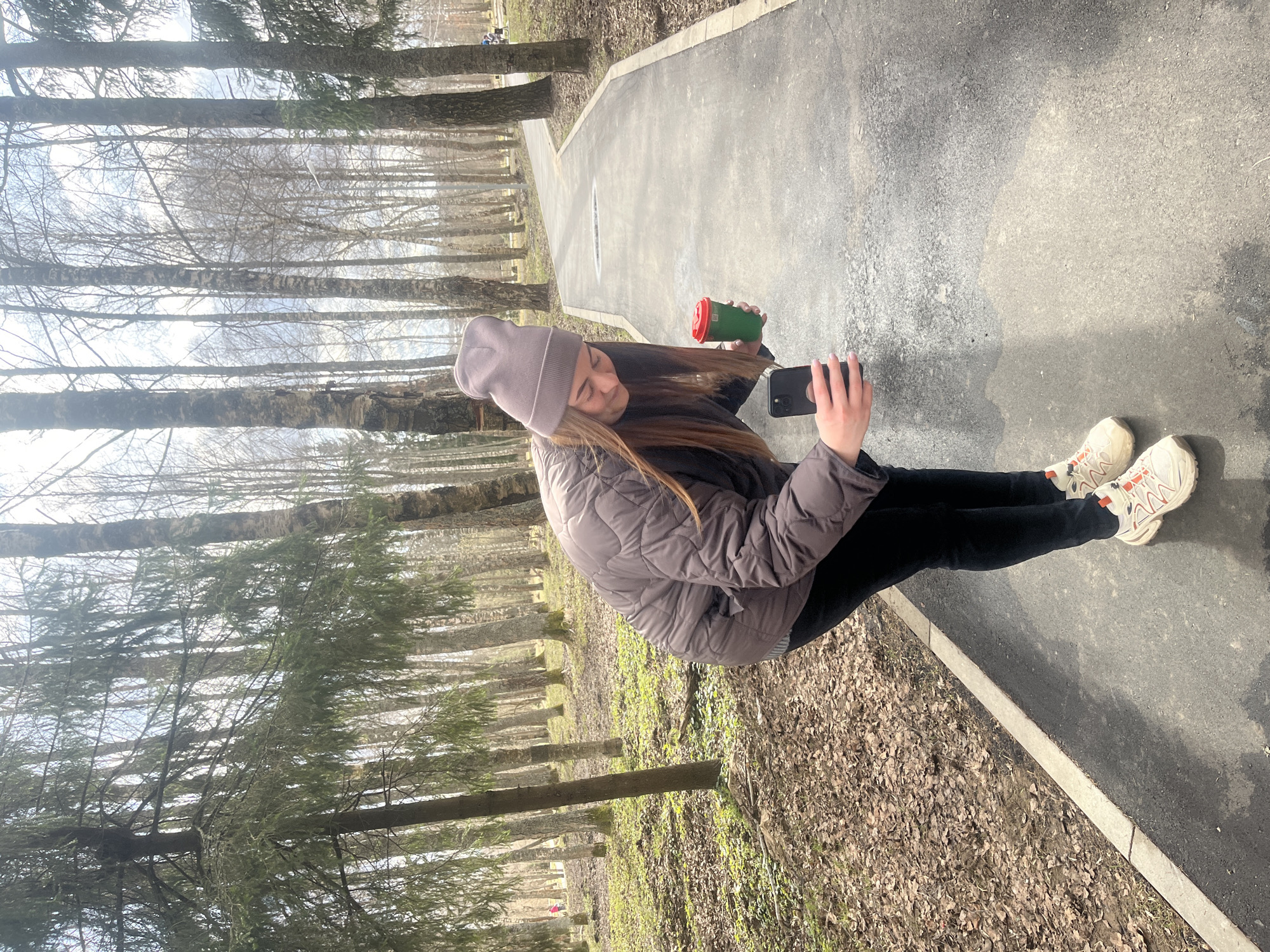 A young woman stands on a gray asphalt path in a park in Vysokovsk, Russia. She is wearing a lilac-colored beanie, a gray quilted jacket, black leggings, and white sneakers with orange accents. She holds a green and red cup in one hand and a black smartphone in the other, seemingly taking a photo or video. The path is bordered on one side by a strip of leaves and ground cover, and on the other by a grove of bare trees. The trees indicate it is likely early spring or late fall. The path appears to be paved and leads into the distance.