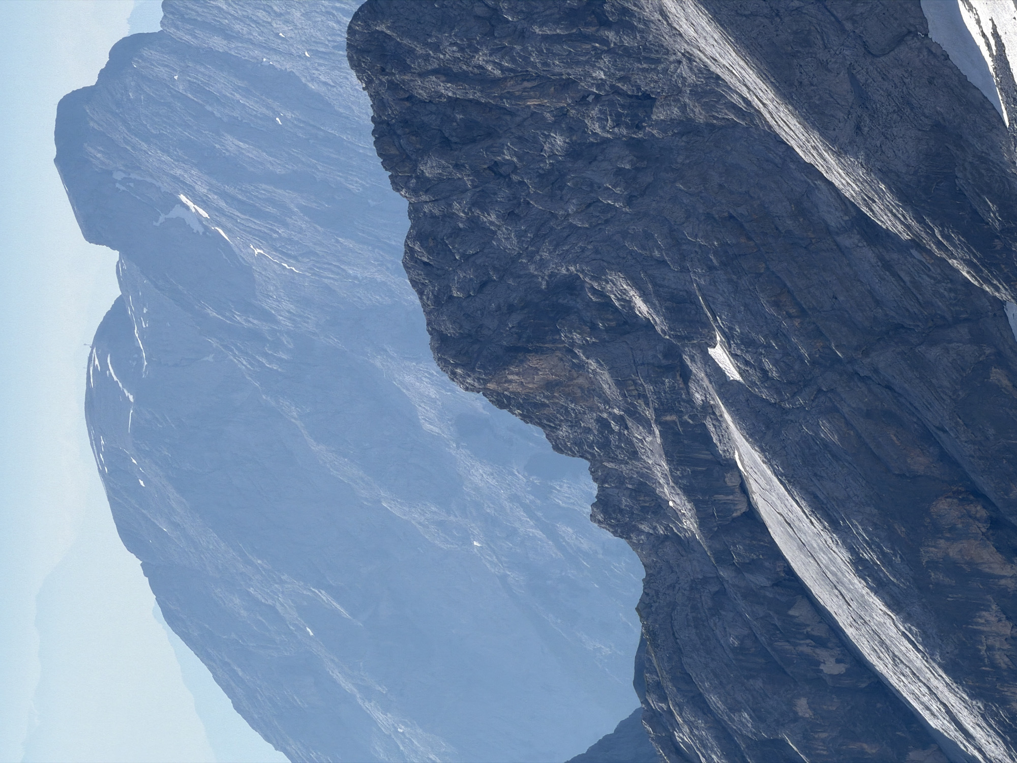 The image shows a layered panorama of mountains, likely in the Bavarian Alps near Garmisch-Partenkirchen, Germany, given the characteristic mountain formations. In the foreground, a dark, rocky mountain face dominates, with distinct striations and crevices. A lighter, scree-covered slope runs diagonally across this mountain. Behind this, a mid-ground mountain range appears in a cooler, bluish hue due to atmospheric perspective, showing patches of snow and rugged terrain. The background features even more distant, hazy mountain peaks, fading into a pale blue sky. The scene is devoid of any human presence or artificial structures, focusing solely on the natural grandeur of the mountainous landscape.