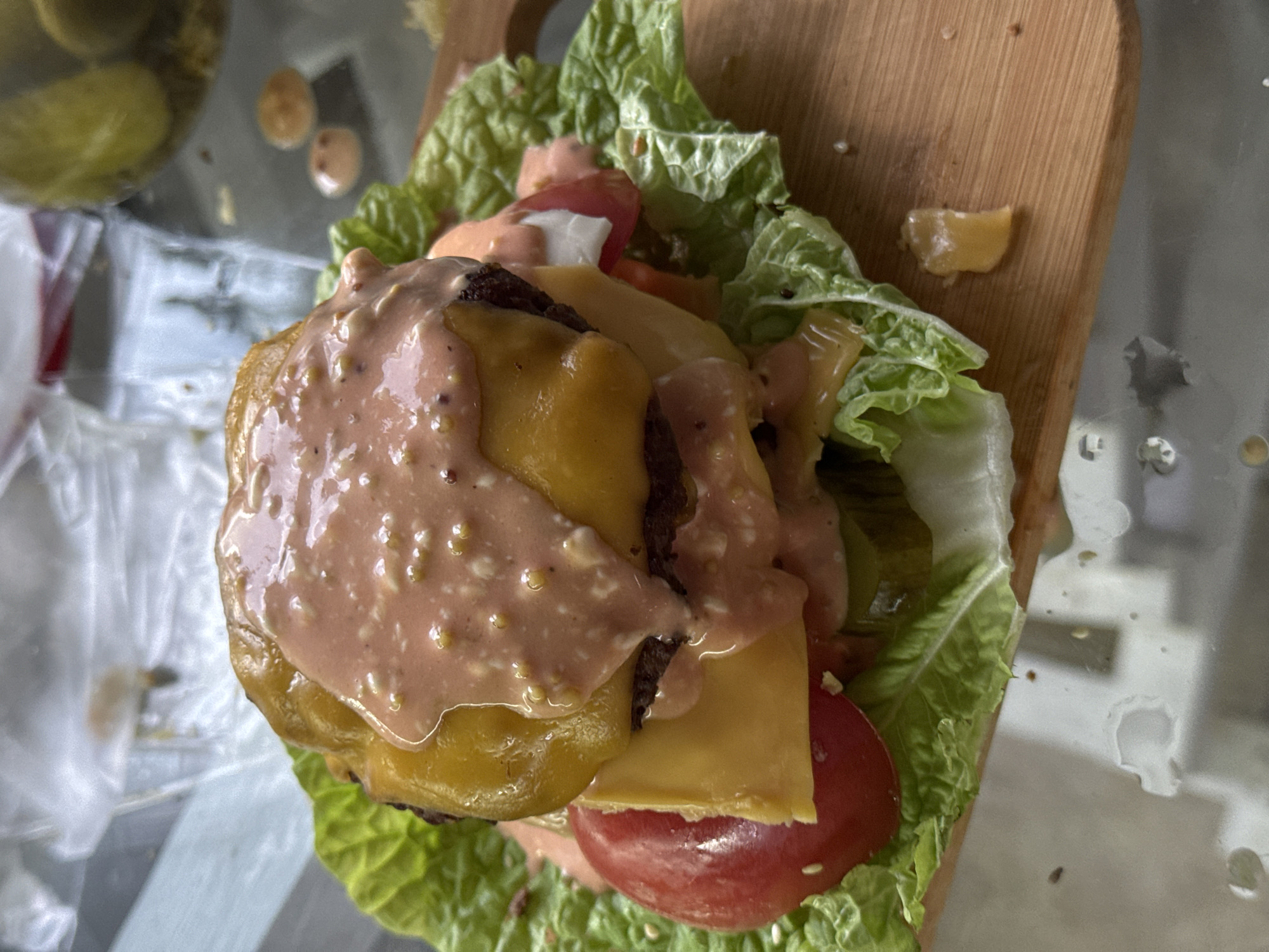 A close-up, overhead shot shows a cheeseburger served on a wooden cutting board. The burger is stacked high with two patties, melted cheese, lettuce, and tomato slices, all covered in a pinkish sauce. The background is out of focus, showing a blurred environment with a hint of a glass surface. The image does not depict any specific location in Ponazyrevo, Russia.
