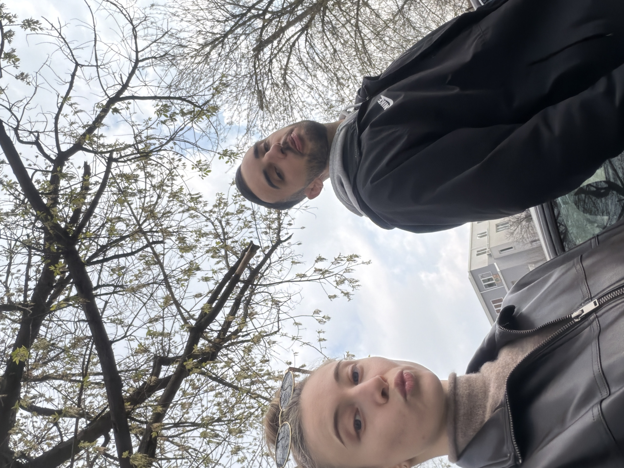 A low-angle, selfie-style photograph depicts two individuals outdoors in Plovdiv, Bulgaria. The female subject, positioned in the lower-left foreground, has light hair pulled back into a bun, with round sunglasses resting on her head. She wears a dark leather-look jacket over a brown turtleneck and poses with pursed lips, looking towards the camera. To her right, the male subject, partially cropped on the right, has dark hair and a beard, wearing a black jacket with a white Nike logo over a grey hooded sweatshirt. He looks directly at the camera with his mouth slightly ajar, exposing a portion of his tongue. The upper half of the frame is filled with a network of dark tree branches, some bare, others displaying nascent light green leaves and catkins, set against an overcast sky with visible patches of lighter blue. In the background, behind the individuals and a dark vehicle partially visible below them, are multi-story urban buildings, including a prominent light-gray structure with multiple windows and visible rooftop apparatus.