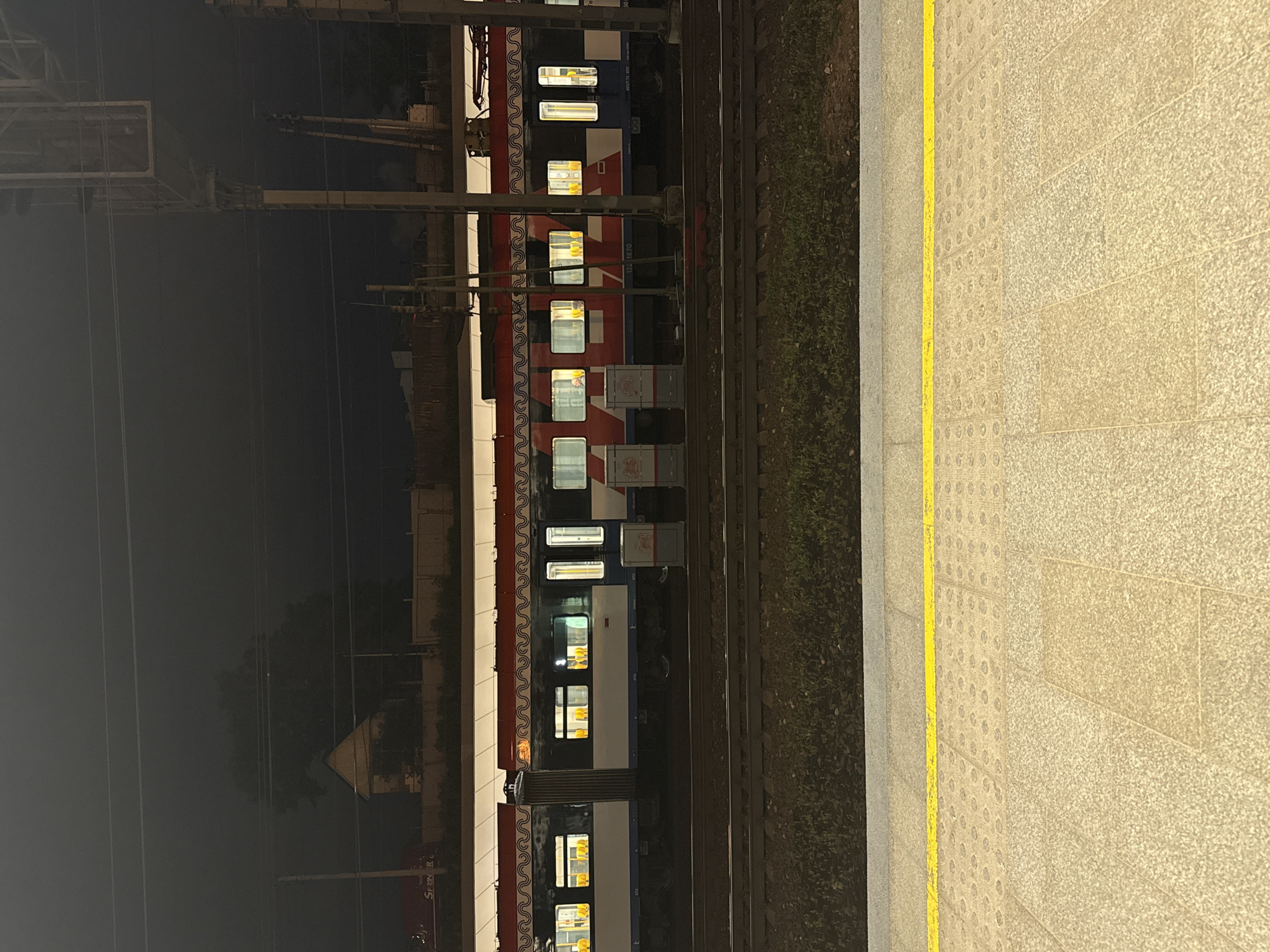 The image depicts a train at a station in Moscow, Russia, at night. The train is red and white, with illuminated windows. It sits on train tracks parallel to the platform. The platform is constructed of light-colored paving stones, marked with a yellow safety line along the edge. The area beyond the tracks and platform is dark, with faint outlines of foliage and structures. Overhead power lines and structural supports are visible above the train. A sign with the word "Sprinter" can be seen on a building in the background.