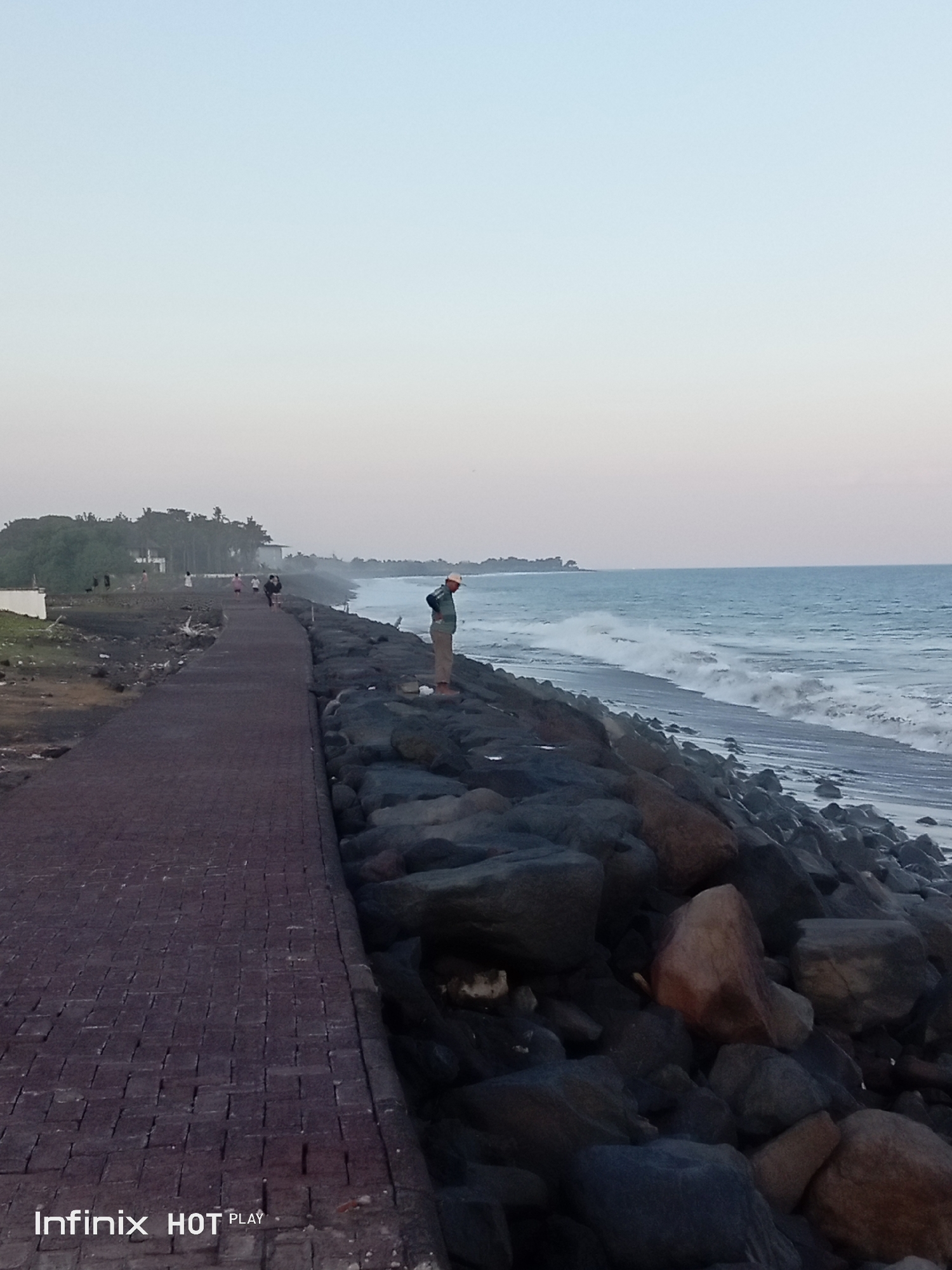 An individual stands on a rocky breakwater in Banjarangkan, Indonesia, facing the ocean. The breakwater is constructed of dark, irregularly shaped stones and borders a paved walkway made of interlocking reddish-brown bricks. The walkway extends into the distance on the left side of the image, curving slightly. The ocean is to the right, with moderate waves crashing onto a dark sandy shore. The sky is a soft gradient of pale blue to a lighter hue, indicating either dawn or dusk. In the background, a line of trees and buildings are visible along the coastline. Several other people are seen walking on the paved pathway further down. The overall scene depicts a coastal promenade with natural and man-made elements.