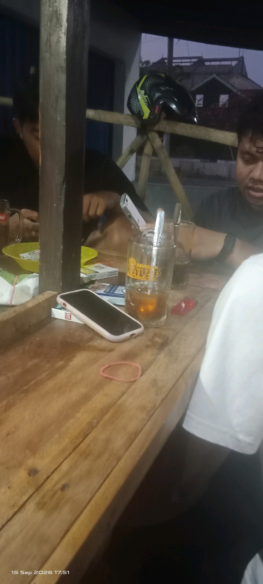 The image depicts an outdoor gathering at night, possibly at a roadside stall or a casual eatery in Merbung, Indonesia, as suggested by the timestamp. The scene is illuminated by artificial lighting, casting shadows on the wooden table and surrounding bamboo structures.
In the foreground, a wooden table is covered with various items. A smartphone with a pink case lies on the table, next to a pack of cigarettes. A pink rubber band is also visible on the table. Further back on the table, a glass contains a amber-colored liquid, likely a beverage, with a metal spoon inside. Next to it is another glass, partially obscured. A yellow plastic bowl with some items, possibly food or snacks, is also present.
Several individuals are present, though their faces are not clearly visible or are out of focus. On the left, a person appears to be looking down at the table. In the middle, a person is holding a smartphone, seemingly interacting with it. On the right, another person is visible from the shoulder up, wearing a dark t-shirt. Their facial features are discernible but somewhat blurred.
In the background, a black and yellow motorcycle helmet rests on a bamboo railing. Beyond that, a building with a dark roof and windows is visible against the dusky sky. The overall ambiance suggests a relaxed, informal social gathering.