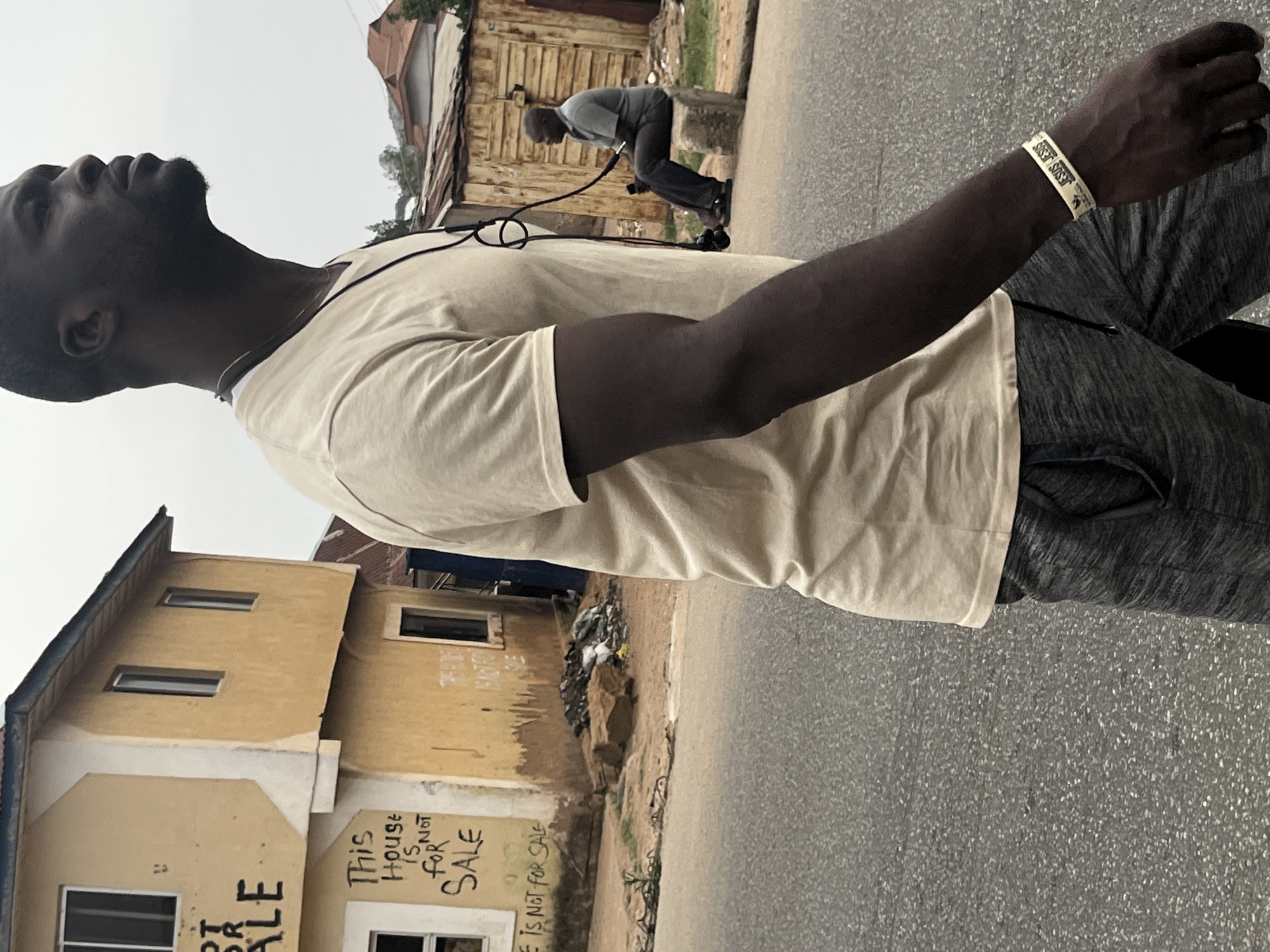 The image shows a man with dark skin in the foreground, wearing a light tan t-shirt, grey pants, and a white wristband. He is looking upwards. Behind him, there is a man bent over, appearing to be working with equipment connected by a hose. In the background, a weathered yellow building has graffiti that reads "This House is Not For Sale". The scene takes place on a paved road in an urban environment. It is believed to be in Jos, Nigeria.