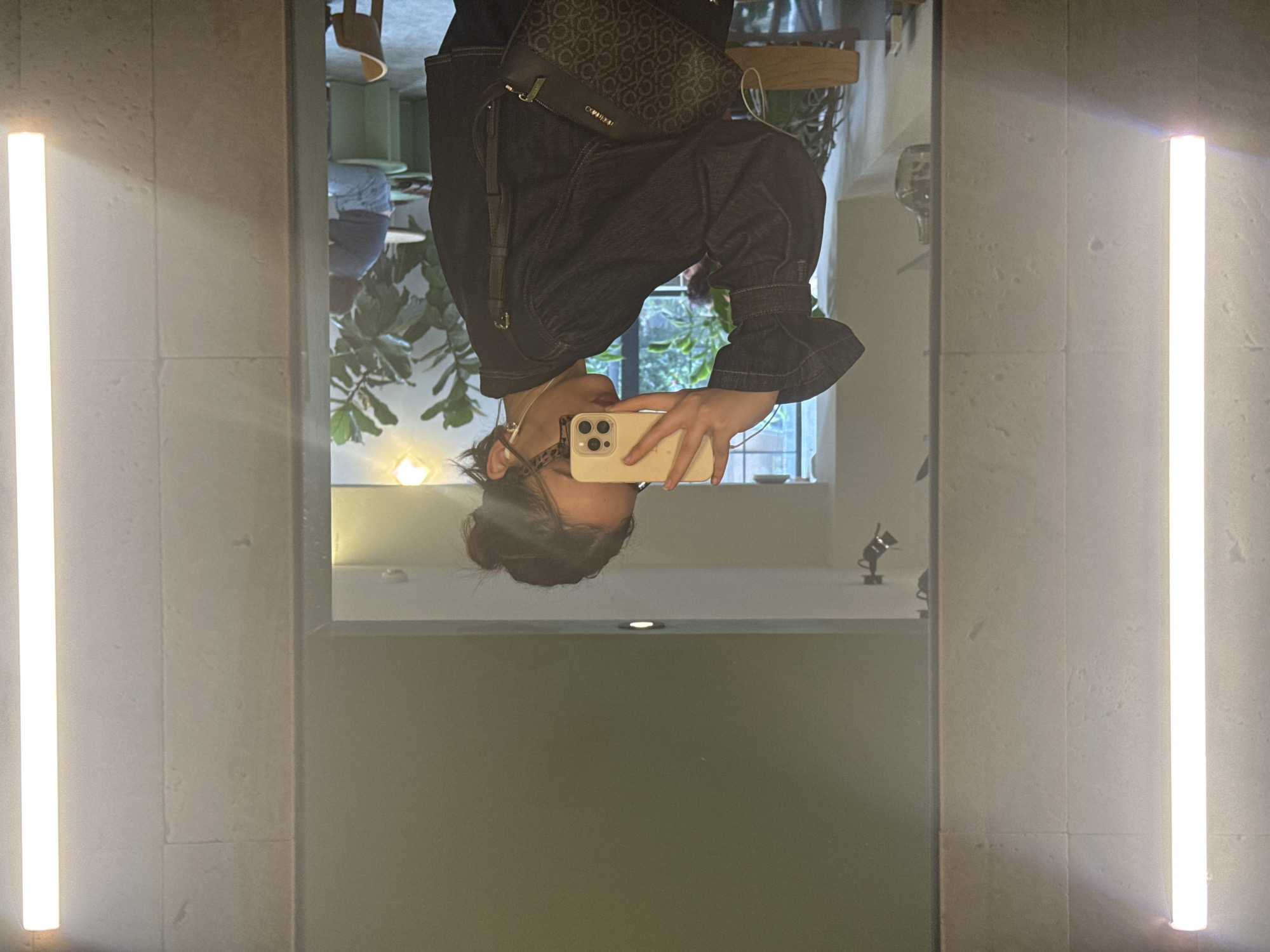 A person, likely female, takes a selfie in a mirror. The reflection shows a dark denim jacket, a Calvin Klein bag, and the person holding a smartphone. The background includes plants and furniture, possibly in a cafe or restaurant setting. The mirror is flanked by two vertical light fixtures. The setting appears indoors. It is not possible to ascertain the location in Yerevan, Armenia, from the information present in this image.