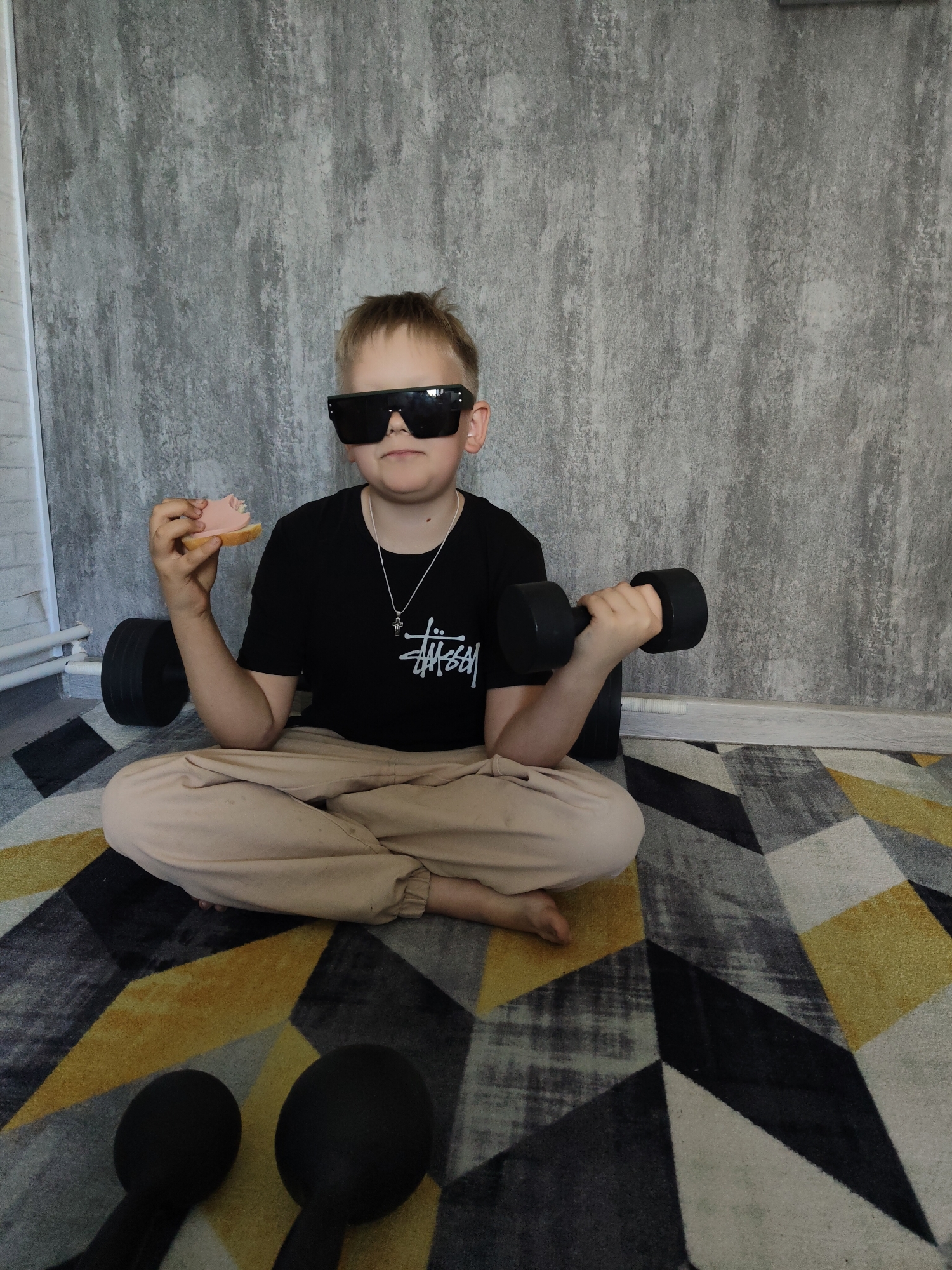 A young person is sitting cross-legged on a geometrically patterned rug with gray, black, white, and yellow sections. The person is wearing large, dark sunglasses, a black t-shirt with a white logo, beige pants, and a silver necklace with a cross pendant. In their left hand, they hold a piece of bread with processed meat on top, and in their right hand, they are lifting a black dumbbell. Two additional black dumbbells are on the rug in front of the person, and another dumbbell is beside them. Behind the person is a wall with a gray textured pattern. The location is Unknown, Unknown.