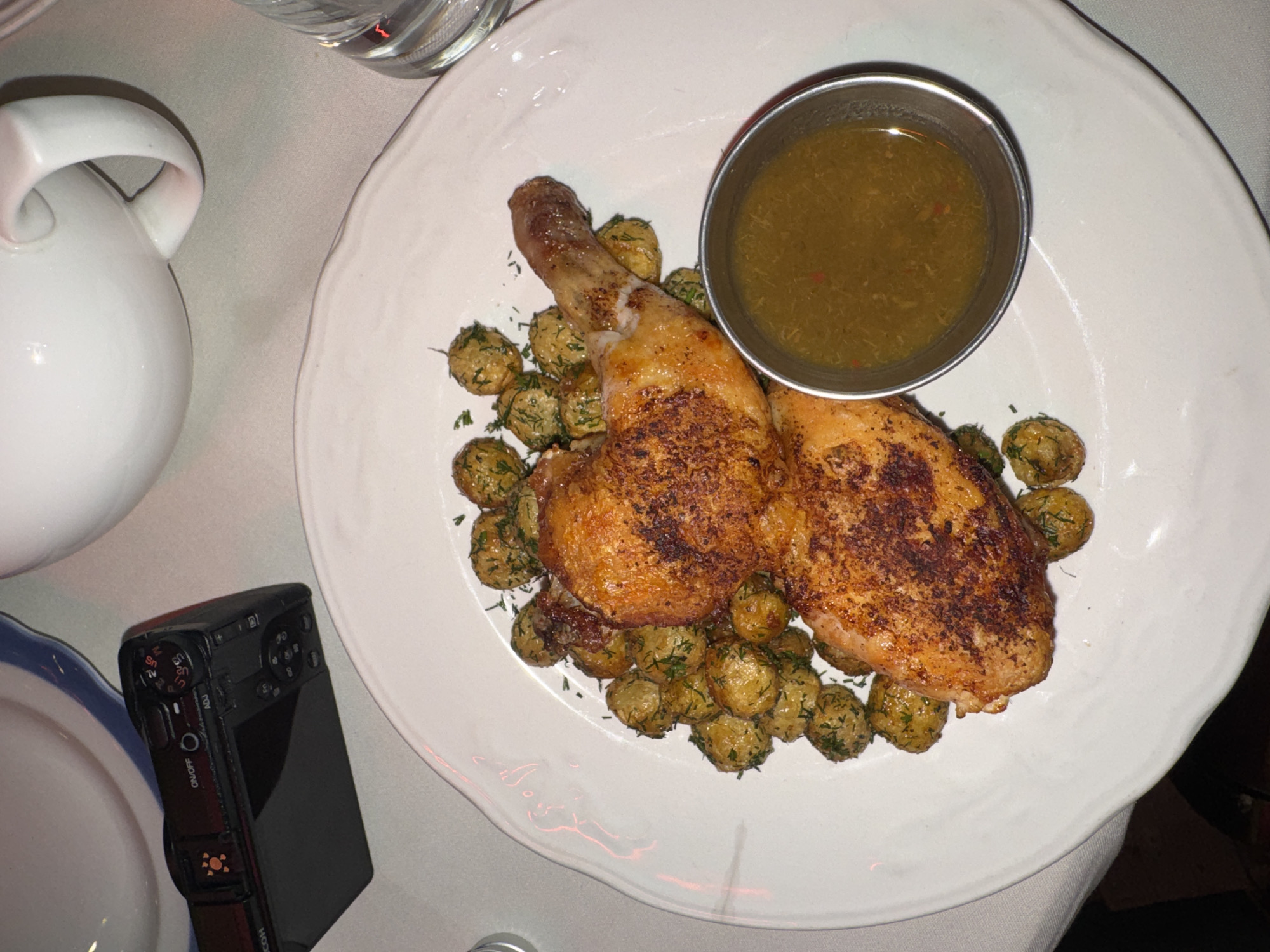 An overhead shot depicts a dinner plate featuring a meal set on a white tablecloth, likely in a restaurant. The main dish consists of two pieces of roasted chicken, appearing to be legs and thighs, with crispy, browned skin. These are surrounded by numerous small, round potatoes, which are garnished with chopped green herbs, likely dill. A small, silver-colored metal cup containing a green-brown sauce, possibly a gravy or vinaigrette, sits on the plate alongside the chicken.
To the upper left of the plate, a white ceramic pitcher with a handle is partially visible, along with the rim of a clear drinking glass. In the lower-left corner of the frame, a black compact digital camera, specifically a Ricoh GR series (identified by visible text "RICOH" and the design), lies on its side next to a partially visible blue and white rimmed plate. Text visible on the camera includes "ON/OFF", "ADJ", and various mode dial settings such as "Sn", "Tv", "Av", "M", "S", "U1", "U2", "U3".
The lighting is dim and warm, suggesting an indoor evening setting. The overall scene captures a meal ready to be eaten. No people are visible. The atmosphere appears calm and focused on the food presentation.