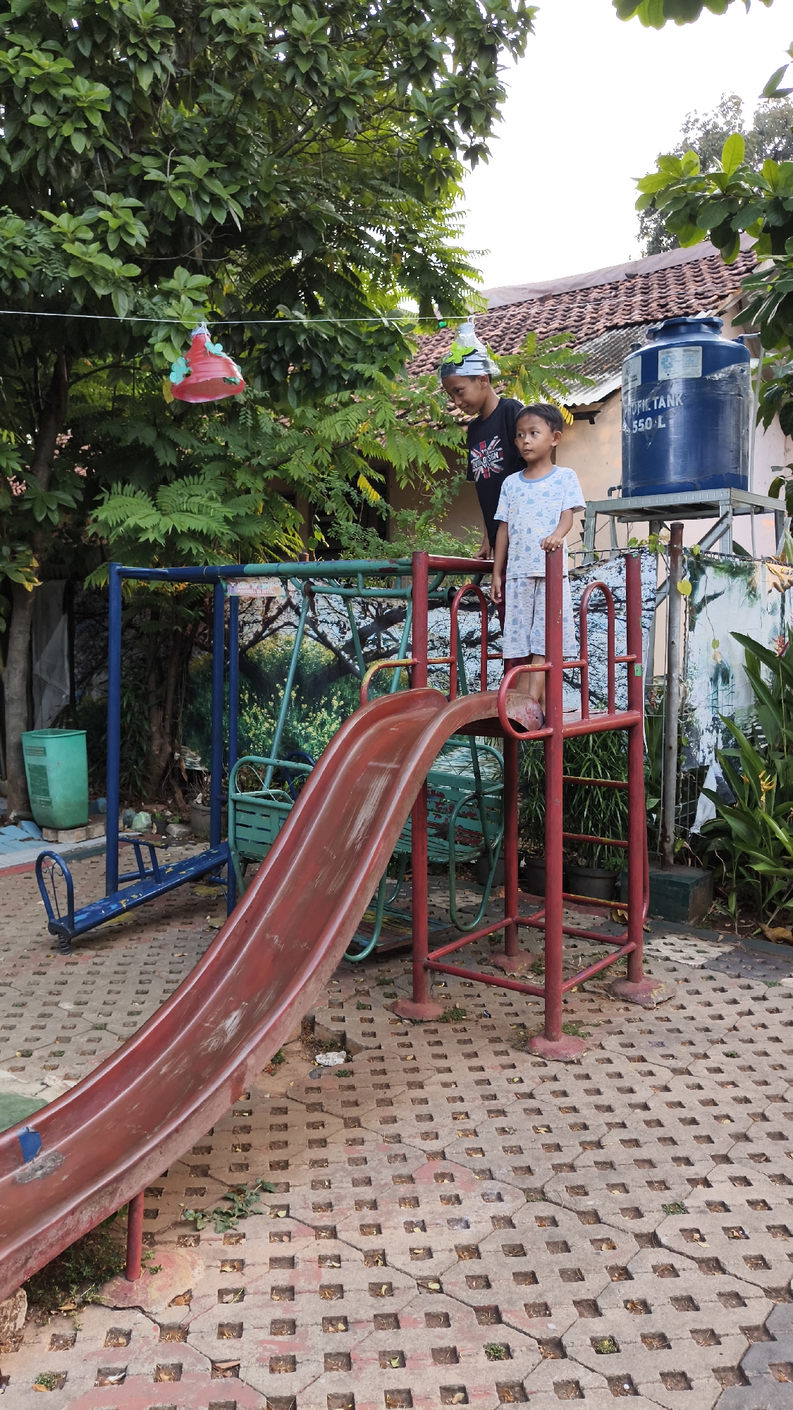 In South Jakarta, Indonesia, two children stand on a playground. The taller child, wearing a black t-shirt and a party hat, looks down at the younger child. The younger child, in a light blue t-shirt and patterned shorts, stands on the top of a red slide, looking towards the right. The playground features a red slide, a blue seesaw, and a green swing set. Behind the playground, a large blue water tank labeled "PROFIL TANK 550-L" is visible, along with trees, a tiled roof, and a textured concrete ground.