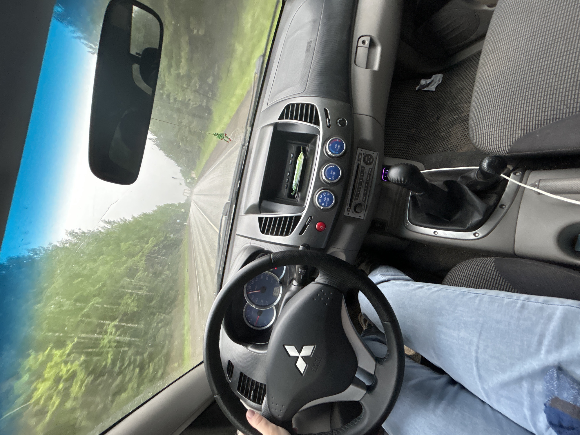 The image shows the interior of a car, likely a Mitsubishi, from the driver's perspective. The dashboard, steering wheel, and center console are visible. The driver is wearing light blue jeans and appears to be holding the steering wheel. Outside the windshield, a road stretches ahead, flanked by green trees. The scene suggests a drive along a road surrounded by vegetation, possibly in a rural or suburban area. The car is in Фоки, Russia. The windshield has some visible damage.