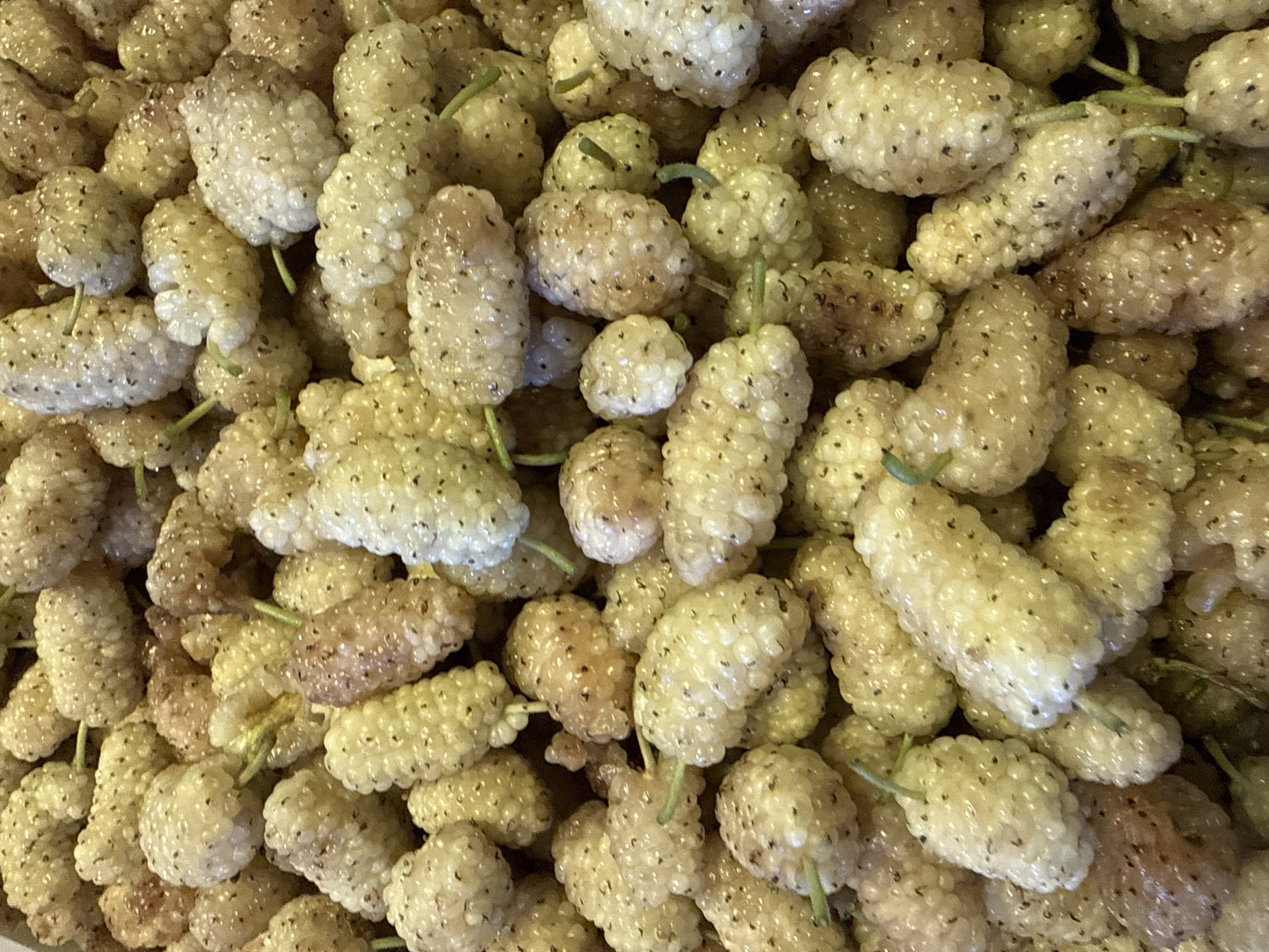 A close-up, overhead shot reveals a dense pile of ripe white mulberries. The berries are elongated and clustered, with visible seeds and tiny green stems. Some berries exhibit a slight pinkish hue, indicating varying stages of ripeness. The texture appears soft and slightly glossy, with light reflecting off their surfaces. The overall impression is one of abundance and freshness. There is no indication of people, actions, interactions, or specific environmental context beyond the berries themselves. The image does not provide any information about the location in the city of Lokbatan, Azerbaijan.