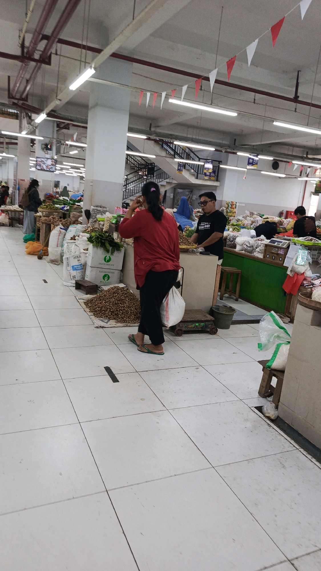 A medium shot captures a busy indoor market scene in Ponorogo, Indonesia. The foreground is dominated by a tiled floor with subtle markings and a black line indicating a boundary. In the mid-ground, a woman in a red top and black pants stands facing away from the camera, holding her hand to her face. She is near a pile of peanuts spread on a mat, flanked by large white sacks. To her right, a man wearing a black t-shirt with text stands behind a counter, appearing to interact with customers. Behind them, various stalls display produce and other goods. The background reveals more market activity, including stairs, overhead lighting, and decorative red and white triangular flags strung across the ceiling. The overall impression is one of a lively commercial environment.
