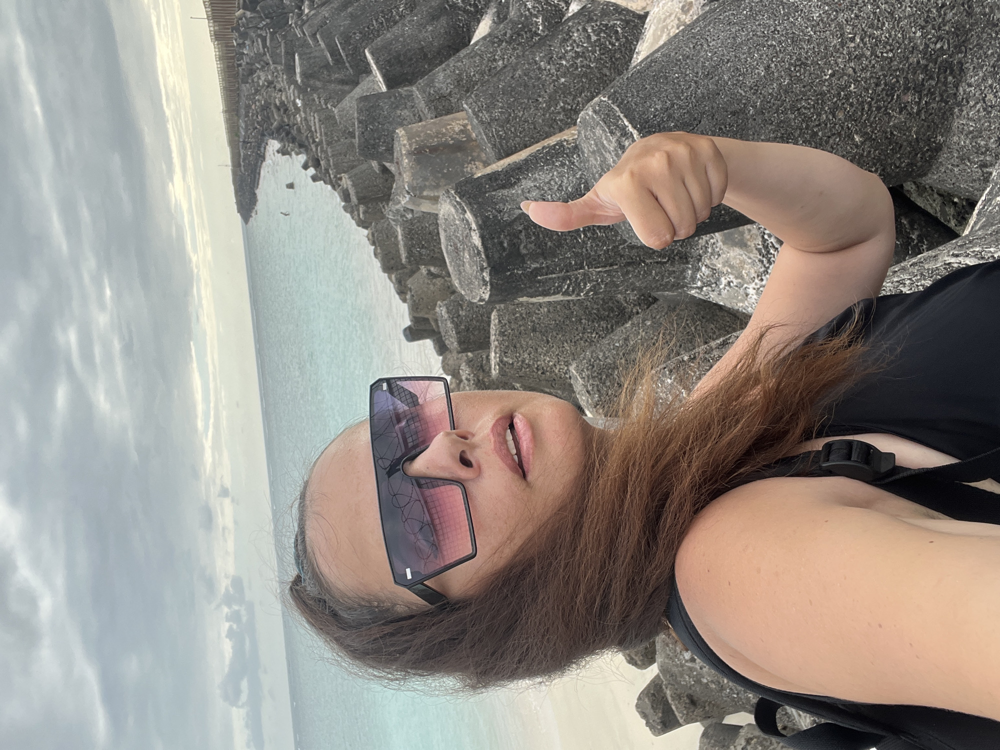 A woman with long brown hair pulled back in a ponytail, wearing oversized sunglasses and a black tank top, takes a selfie on a rocky shoreline. She is giving a thumbs-up with her right hand. Behind her, the turquoise ocean meets a sandy beach, and a breakwater made of large concrete blocks stretches into the distance. The sky is overcast with dramatic clouds. The scene appears to be the beach in Kuta, Bali, Indonesia, known for its surf breaks and tourist attractions.