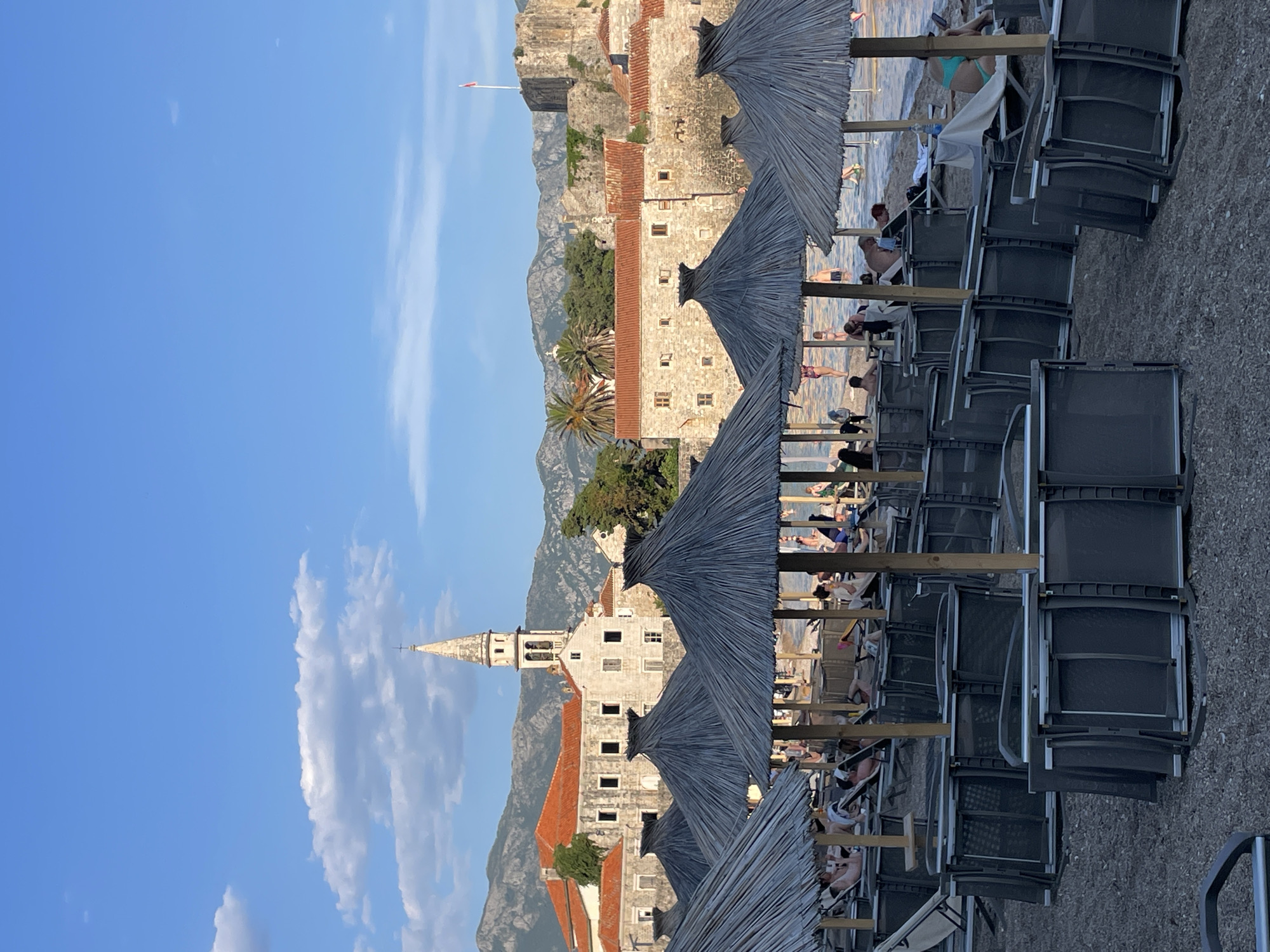 The image depicts a view of the Mogren beach in Budva, Montenegro, with the Old Town visible in the background. The foreground is dominated by rows of empty lounge chairs stacked neatly and thatched umbrellas providing shade. Further back, a small number of beachgoers are visible relaxing on loungers by the water's edge. The historic Old Town of Budva, characterized by its stone buildings with red-tiled roofs and a prominent church tower, is situated behind the beach. Mountains form the backdrop to the scene. The sky is a clear blue with scattered white clouds. The lighting suggests it is late afternoon or early evening.
