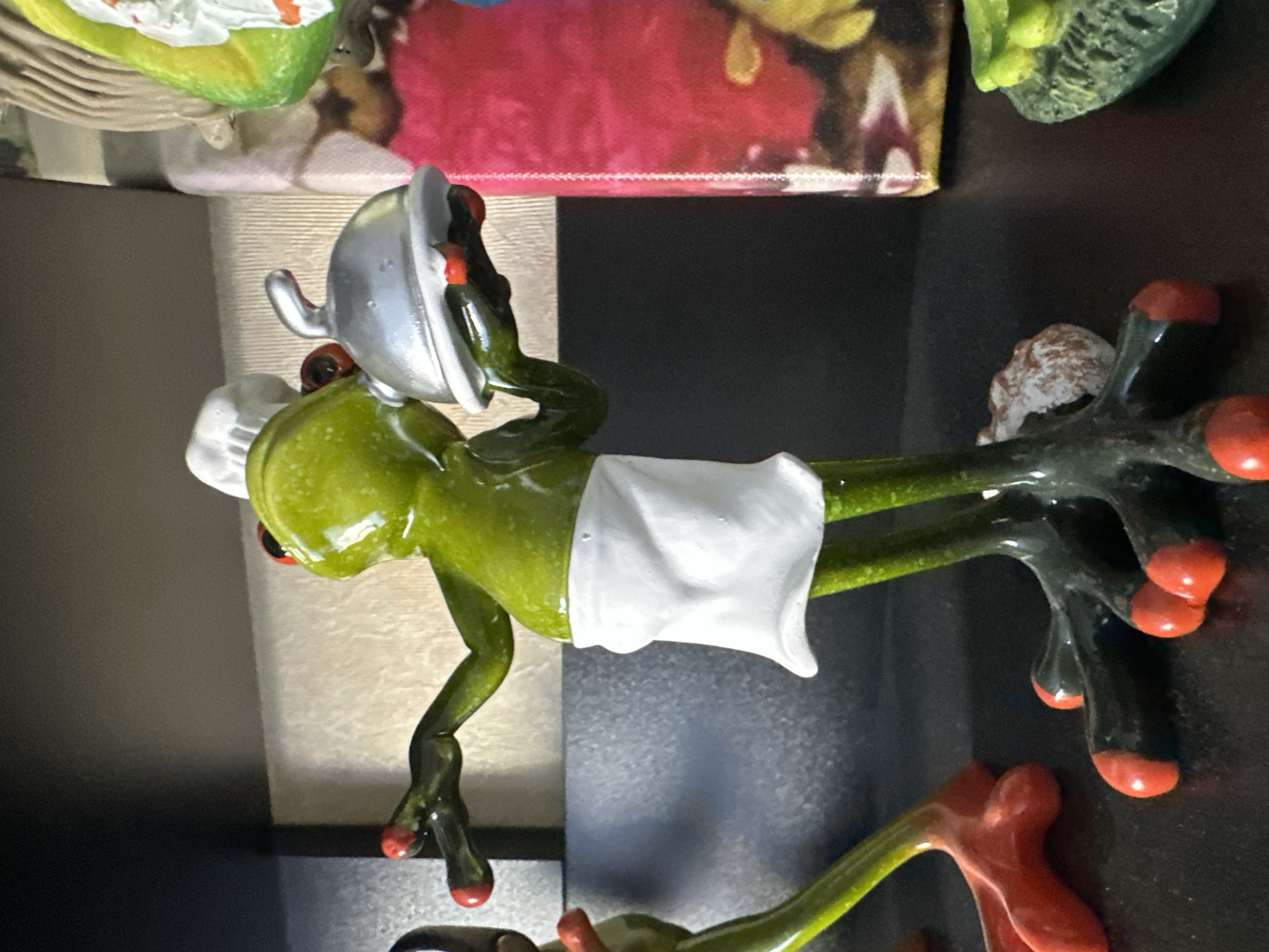 The image shows a close-up, eye-level shot of several small, decorative figurines on a dark, reflective surface, possibly a shelf. The main subject is a green frog figurine dressed as a chef, wearing a white chef's hat and apron. It is holding a silver serving cloche. To the left of the chef frog, another green frog figurine is visible, with red feet. In the background, out of focus, are colorful elements, including a pink, abstract pattern and a green object that appears to be another figurine. The lighting is somewhat dim, with reflections on the dark surface. The scene is static and does not depict any specific activity beyond the posed nature of the figurines. There is no information in the image to suggest a location in Podolsk, Russia.