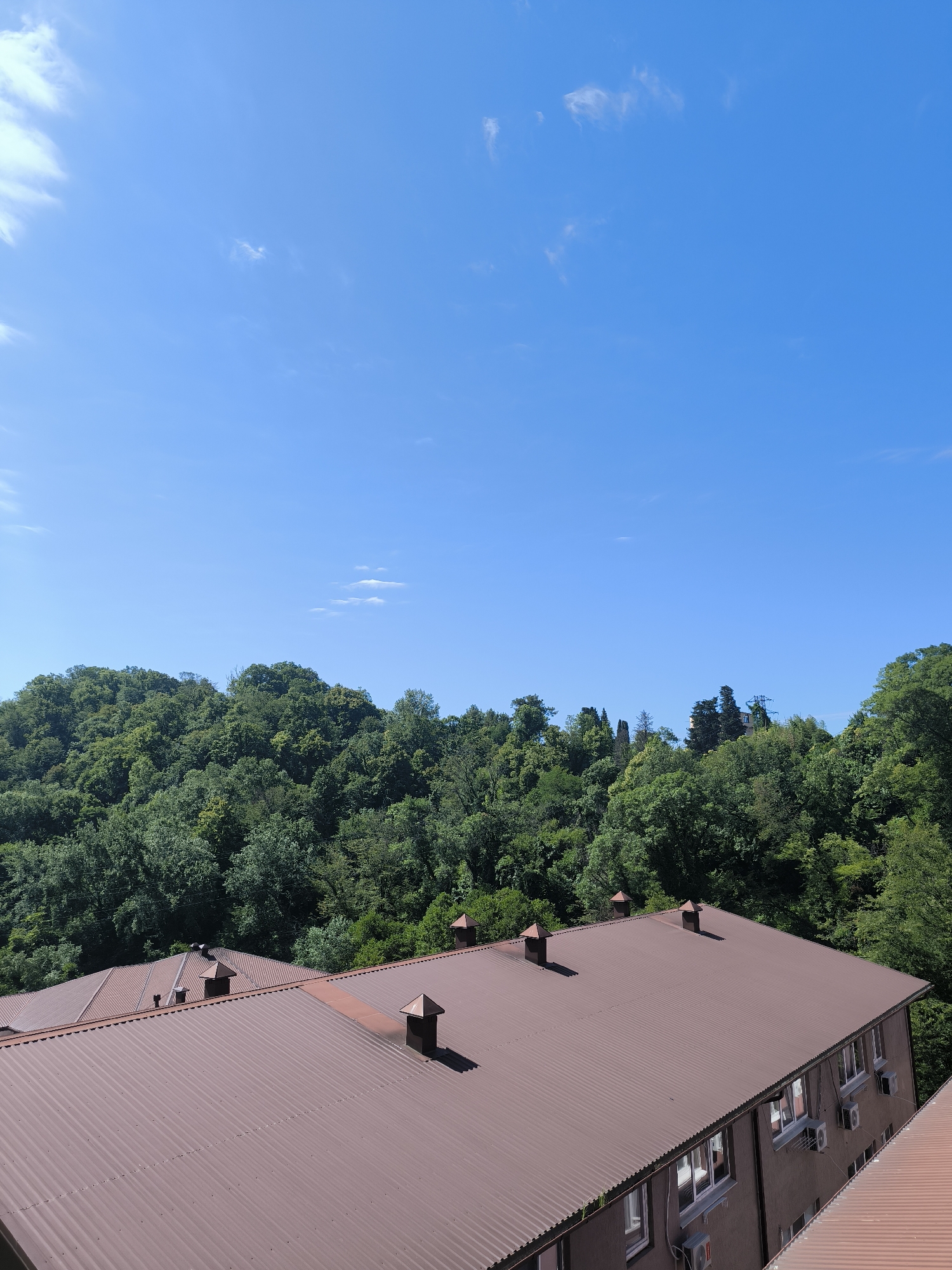 The image captures a clear blue sky with scattered wispy clouds above a dense, verdant forest blanketing rolling hills. In the foreground, two adjacent buildings with corrugated brown metal roofs are visible. The roofs feature several square, dark brown chimneys with pyramidal caps. One building, on the right, has a facade with multiple windows and air conditioning units mounted on the exterior walls. The scene suggests a location in Sochi, Russia, likely an area with residential buildings nestled against a natural, forested landscape. The lighting indicates it is daytime, with the sun casting shadows from the chimneys onto the roof.