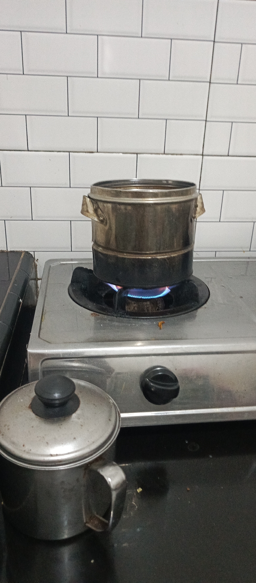 In a kitchen in Arjasa, Indonesia, a stainless steel stove with a single burner is in operation, as indicated by the visible blue flames beneath a multi-tiered stainless steel pot. The pot appears to be a steamer or similar cooking vessel, with two distinct levels visible. To the left of the stove, a smaller stainless steel pot with a lid and handle is placed on a dark countertop. The background consists of a white tiled wall with dark grout lines. The scene suggests food preparation is underway.