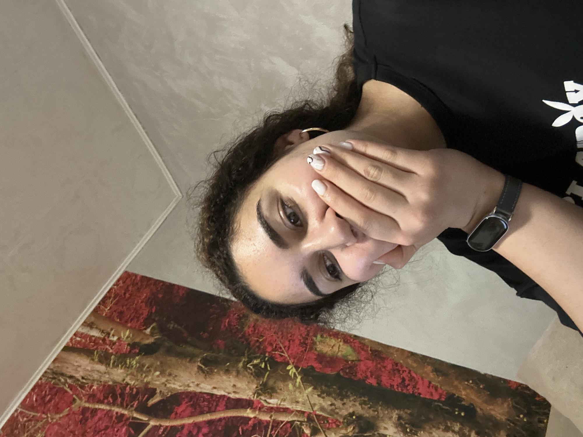 A woman is shown in an indoor setting. The woman has curly brown hair, brown eyes, and manicured nails. She is wearing a black shirt and a wristwatch. She is holding her hand near her face. Behind the woman is a wall, which is partially covered by a picture featuring reddish-brown imagery and tree branches. The photograph does not provide enough information to place the scene within the city of Burgas, Bulgaria.