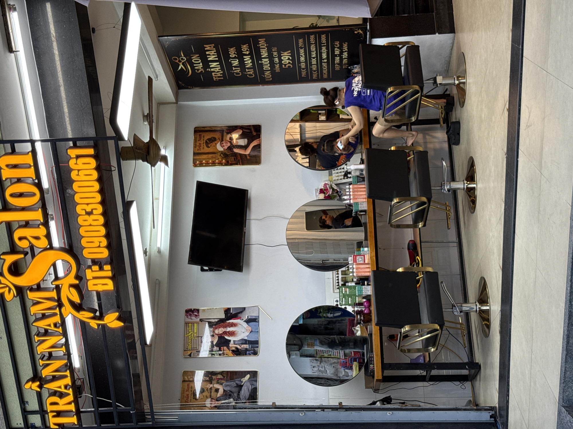 The image depicts the storefront of a salon named "TRÂN NAM SALON" in Nha Trang, Vietnam. A large, illuminated sign above the entrance reads "TRẦN NAM SALON" in orange lettering, with a phone number "ĐT: 0908300661" below it. Inside the salon, there are several styling chairs, mirrors, and a counter stocked with hair products. A television screen is mounted on the wall. Two posters advertising hair services are displayed on the wall to the left. A woman in a blue shirt is seated in a styling chair, appearing to be receiving a haircut from another person whose back is to the camera. The floor is tiled. The overall impression is of a busy and well-equipped hair salon.