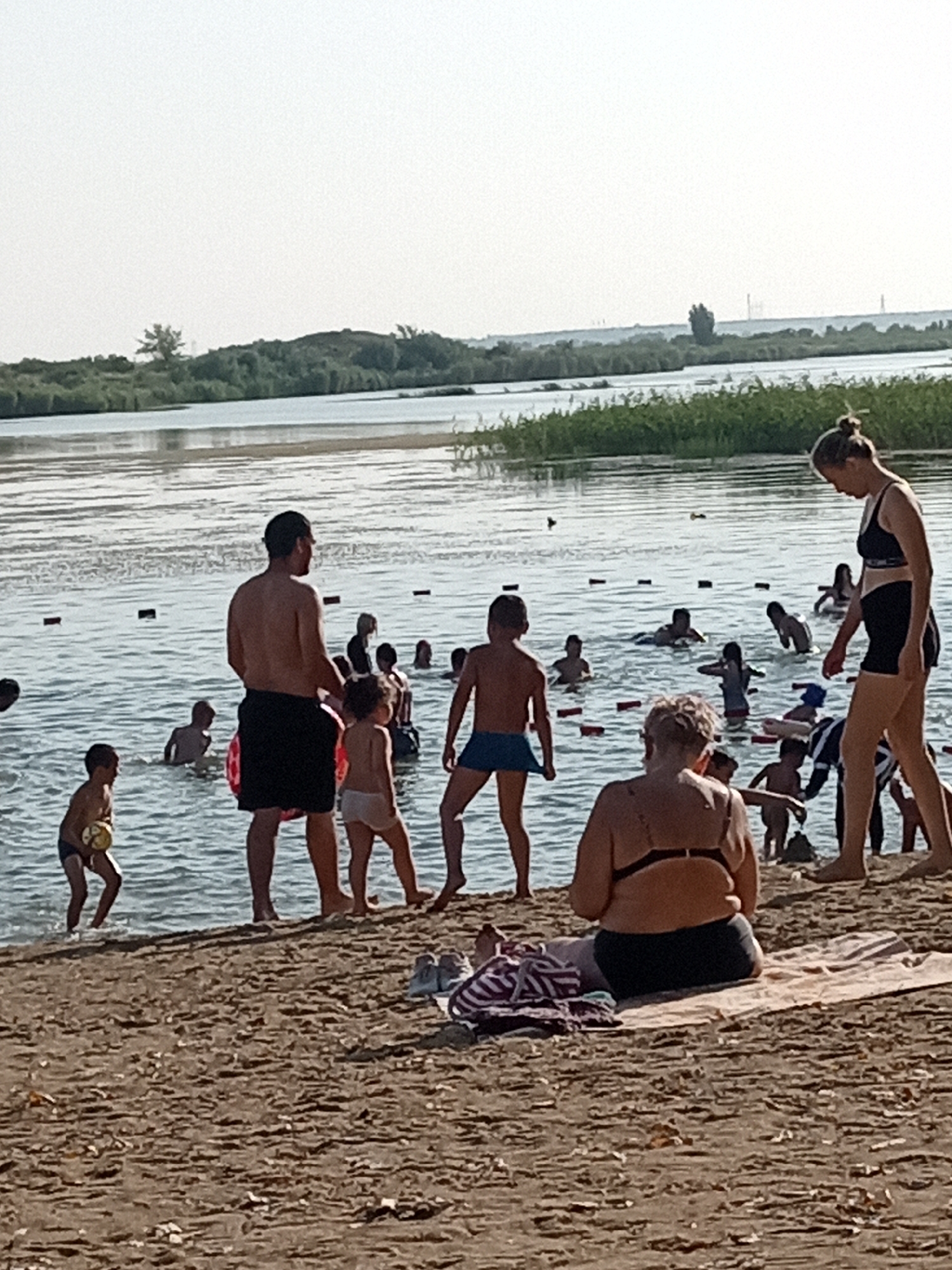 A group of people are swimming and relaxing on the sandy shore of a body of water, likely a lake or river, near Rudny, Kazakhstan. The scene is captured during daylight hours, with the sun casting a bright light. In the foreground, a person wearing a black bikini top and black bottoms sits on a towel on the sand, facing away from the camera and towards the water. To the left of this person, another individual in a black bikini top and black bottoms stands on the sand.
In the water, numerous individuals are visible. A man in black swim shorts stands in the foreground, facing away from the camera. To his left, a child in blue swim shorts holds a yellow ball. Other swimmers are scattered throughout the water, some of them children. The water has a reddish-brown hue, and there are buoys or markers visible in the water.
The background shows a shoreline with reeds and vegetation, and further in the distance, there is a hazy horizon with what appear to be industrial structures or power lines. The sky is overcast or hazy. The overall impression is a casual summer day at a public swimming area.