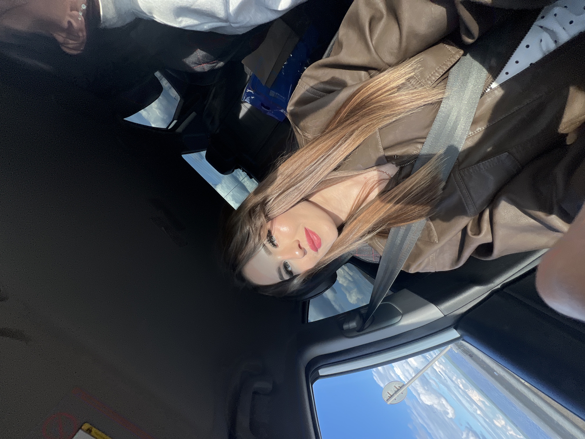 A woman with brown hair and a brown leather jacket is in a car wearing a seatbelt. The scene is an interior car shot, with the sky visible through the car windows. In the lower right window, a street sign is visible. Based on this sign, the location is likely Chaikovsky, Russia.