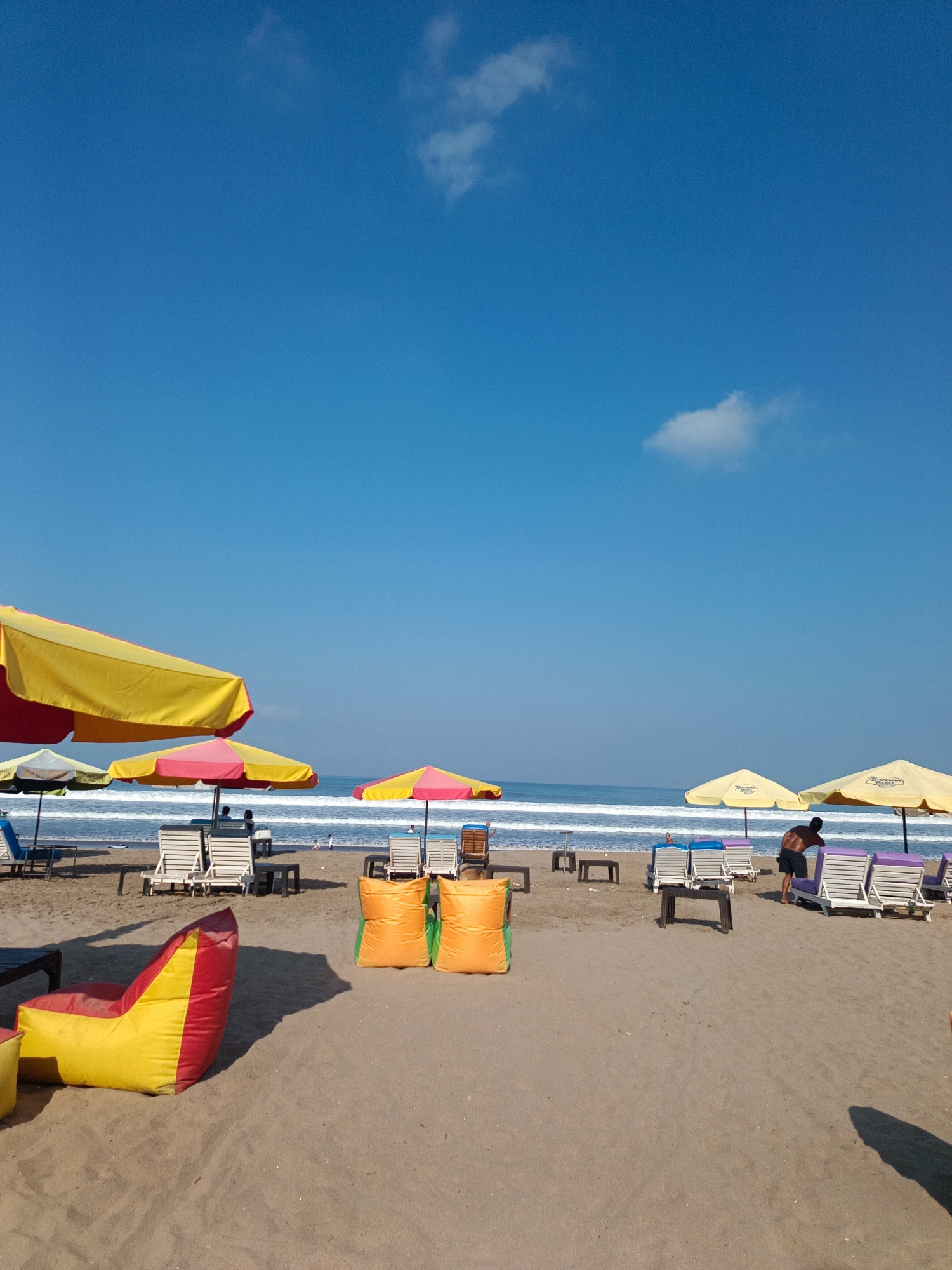 This image captures a beach scene in Kuta, Indonesia. The sand is a light brown, leading to the ocean where gentle waves are breaking. The sky is a clear, bright blue with a few wispy clouds. In the foreground, there are several beach chairs and umbrellas set up. Two large orange beanbag chairs are prominently placed in the center. To the left, a red and yellow beanbag chair is visible. Further back, several white lounge chairs are arranged under colorful umbrellas, some of which are yellow and pink. A few people are present, including one individual wearing black shorts who appears to be adjusting a lounge chair near some yellow umbrellas. In the distance, the ocean stretches out, with small figures of people visible near the water's edge. The overall impression is a sunny day on a well-equipped beach.