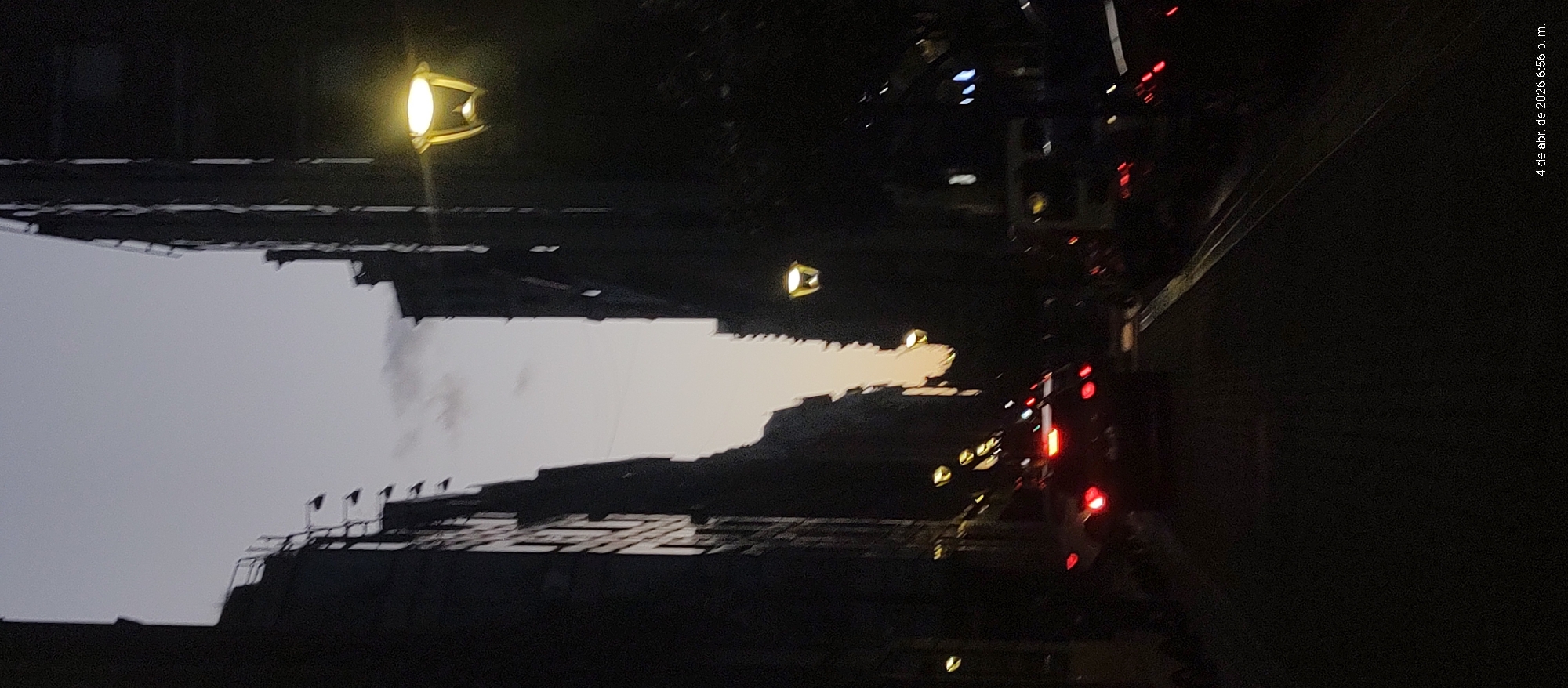 The image is a nighttime shot of a street in Buenos Aires, Argentina. Buildings line both sides of the street, punctuated by street lamps. Several cars can be seen on the road, their headlights and taillights visible. The sky appears to be twilight. A time and date stamp in the upper right corner reads "4 de abr. de 2026 6:56 p.m."