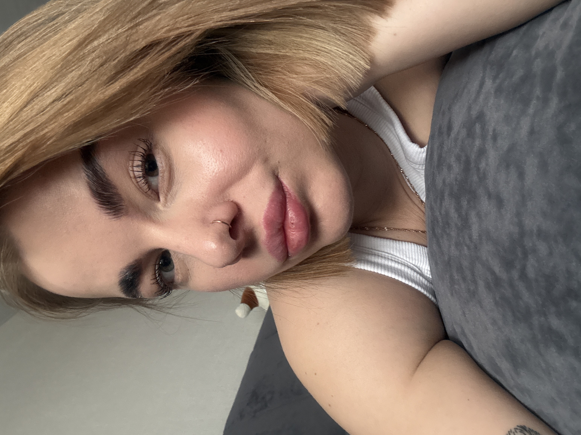 A blonde woman is seen in a close-up, angled shot. She is resting against a grey cushion, wearing a white tank top and a gold necklace. She has a piercing in her left nostril. The photograph was taken indoors. No other details indicate that this photograph was taken in Chaikovsky, Russia.