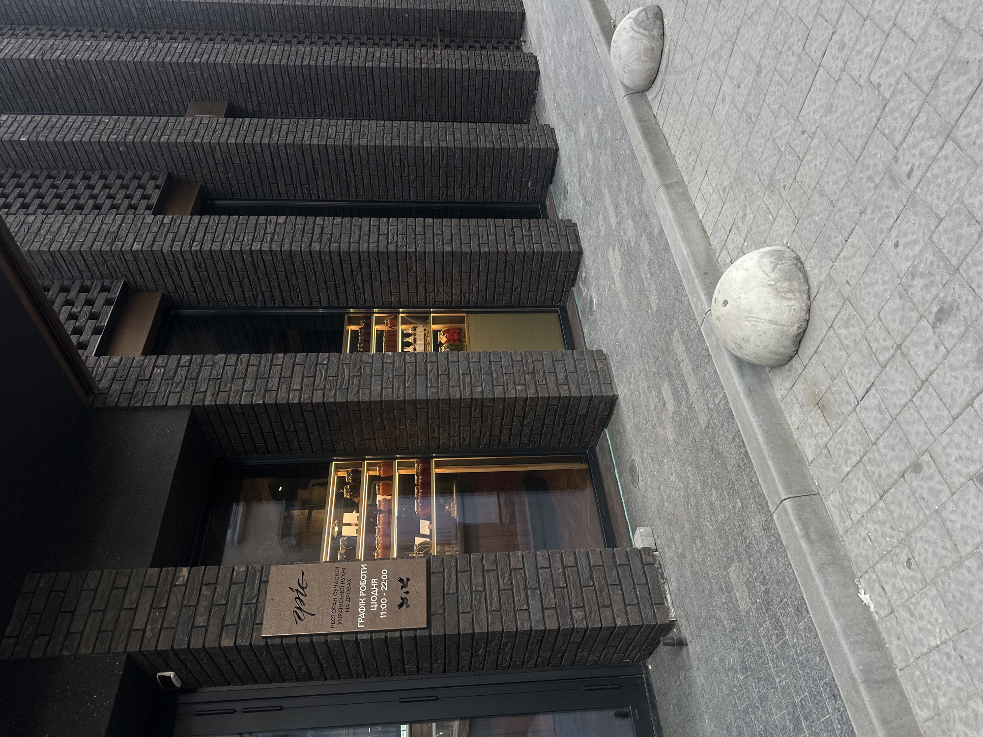 The image shows the exterior of a building in Ternopil, Ukraine. The building has a modern design with dark brickwork. The facade is characterized by stepped-out sections creating a layered effect. There are windows with displays of goods or decorations visible inside. A sign on the building reads "Ресторан сучасної української кухні на дровах", translated as "Restaurant of modern Ukrainian cuisine on firewood," and includes operating hours, "Графік роботи щодня 11:00 - 22:00", or "Operating hours daily 11:00 - 22:00". The sidewalk adjacent to the building is paved with gray interlocking concrete pavers, and a row of stone bollards is placed along the curb. There is no human activity visible in the frame.
