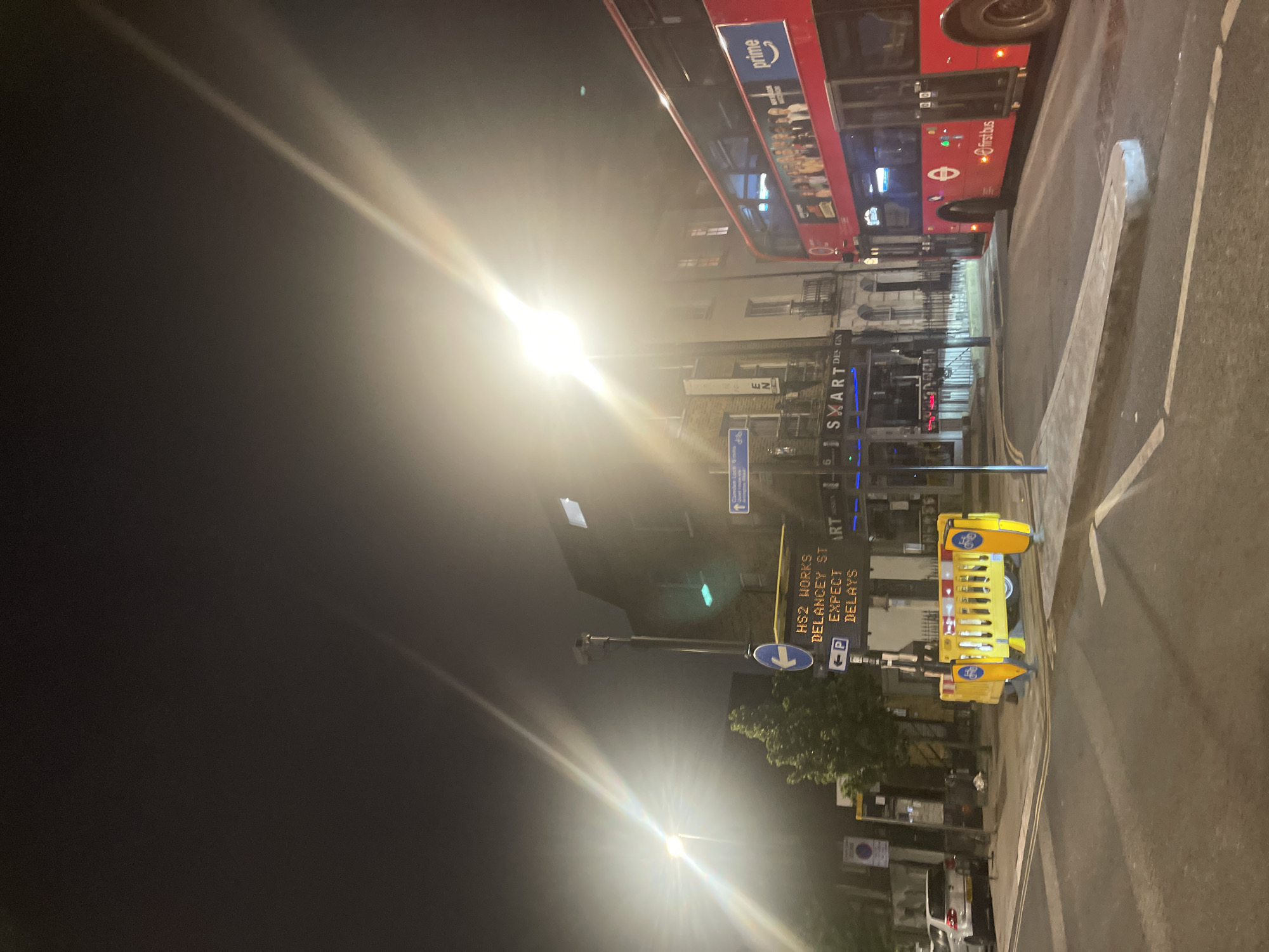 The image shows a street scene at night in London, United Kingdom. A red double-decker bus is present in the top portion of the frame, featuring advertisements. Street signage indicates "HS2 WORKS DELANCEY ST EXPECT DELAYS" and includes a parking symbol. A yellow barrier for cyclists is present. The street is illuminated by streetlights, creating light flares in the image. Buildings line the street, and a sign for "SXART DES GN" is visible. A First Bus vehicle is present. The image also includes a sign for "prime".