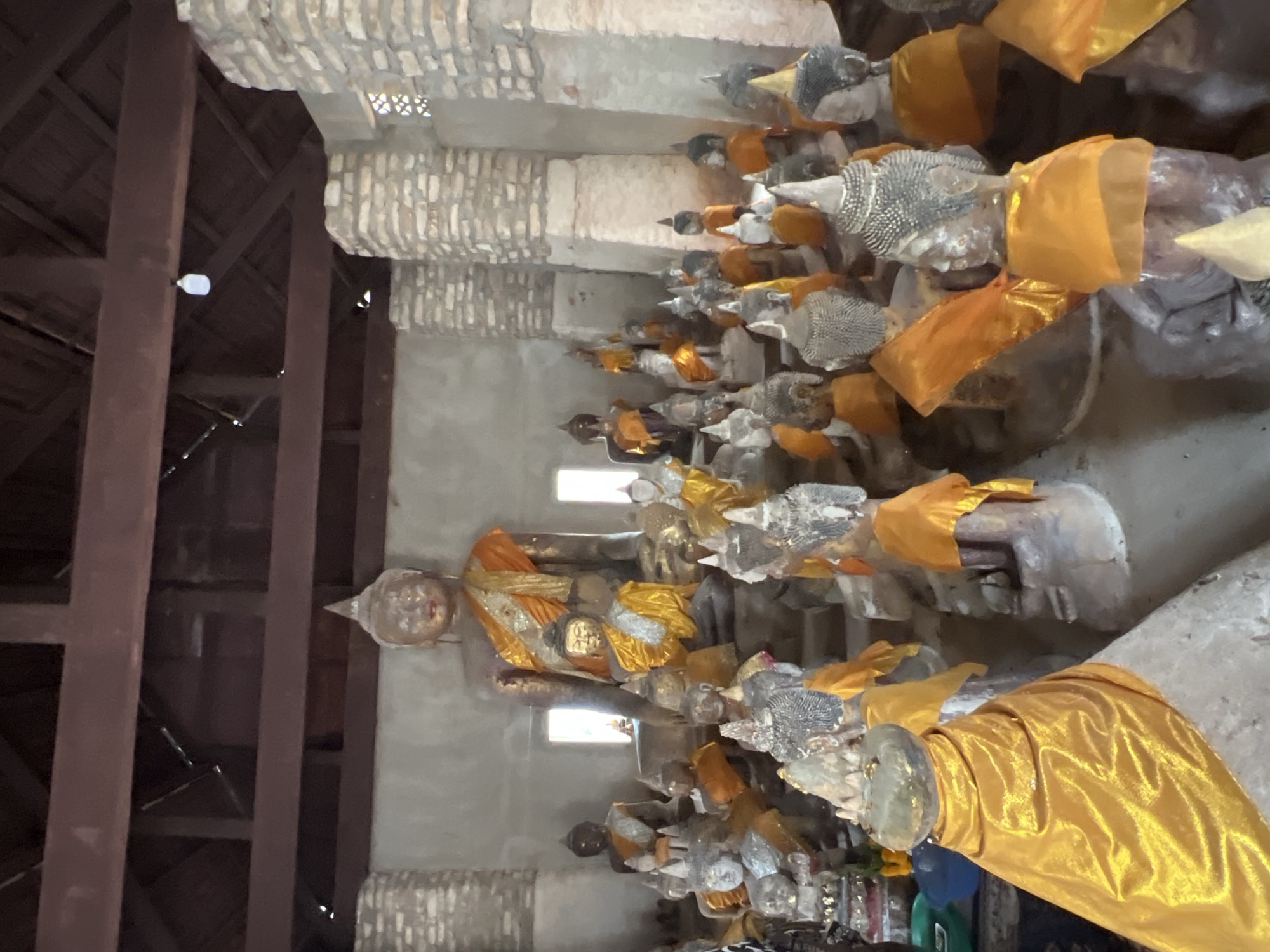 The image shows a collection of Buddha statues in a temple setting, likely in Ko Samui District, Thailand. The statues vary in size, with a large central figure dominating the scene. Many of the statues are adorned with orange and yellow sashes. The architecture of the temple is visible, with rough-hewn stone pillars and a wooden ceiling. The lighting suggests it is daytime, with natural light filtering in from windows or openings. The overall impression is one of a sacred and serene space dedicated to Buddhist worship.
