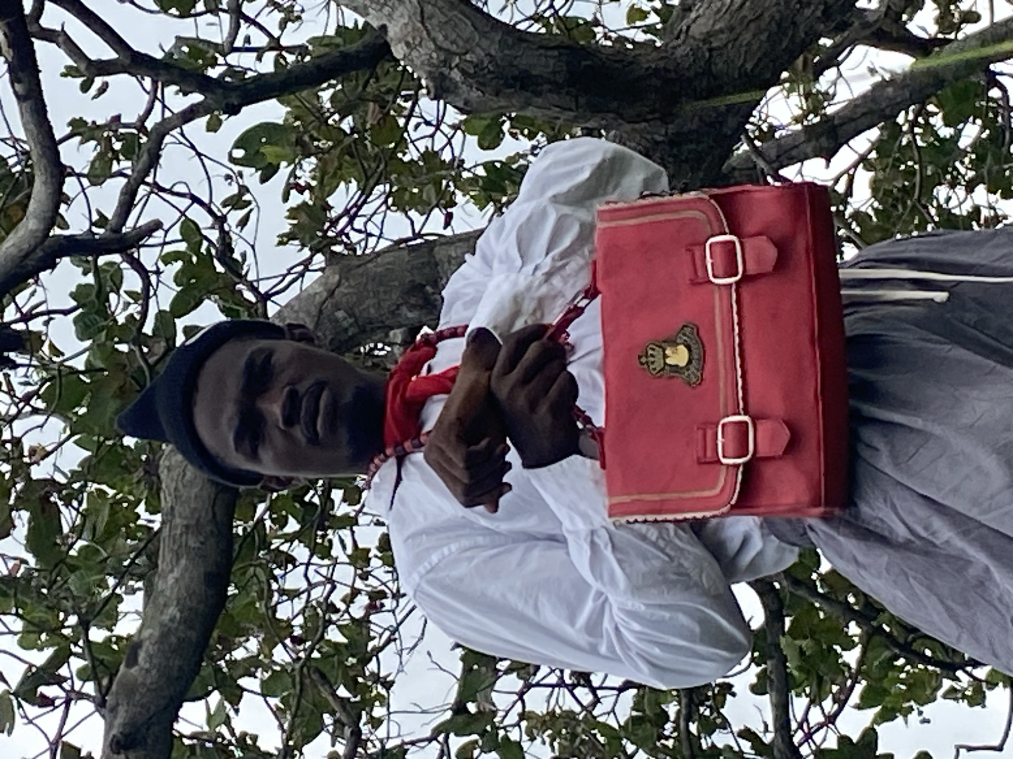 In the city of Ufu Esioni, Nigeria, a person with dark skin, wearing a black conical hat, a white collared shirt with a red scarf tied around their neck, and a grey skirt or trousers is positioned outdoors, partially obscured by the branches of a tree. The person is holding a bright red handbag with gold trim and a crest on its front. The background consists of lush green foliage and a cloudy sky.