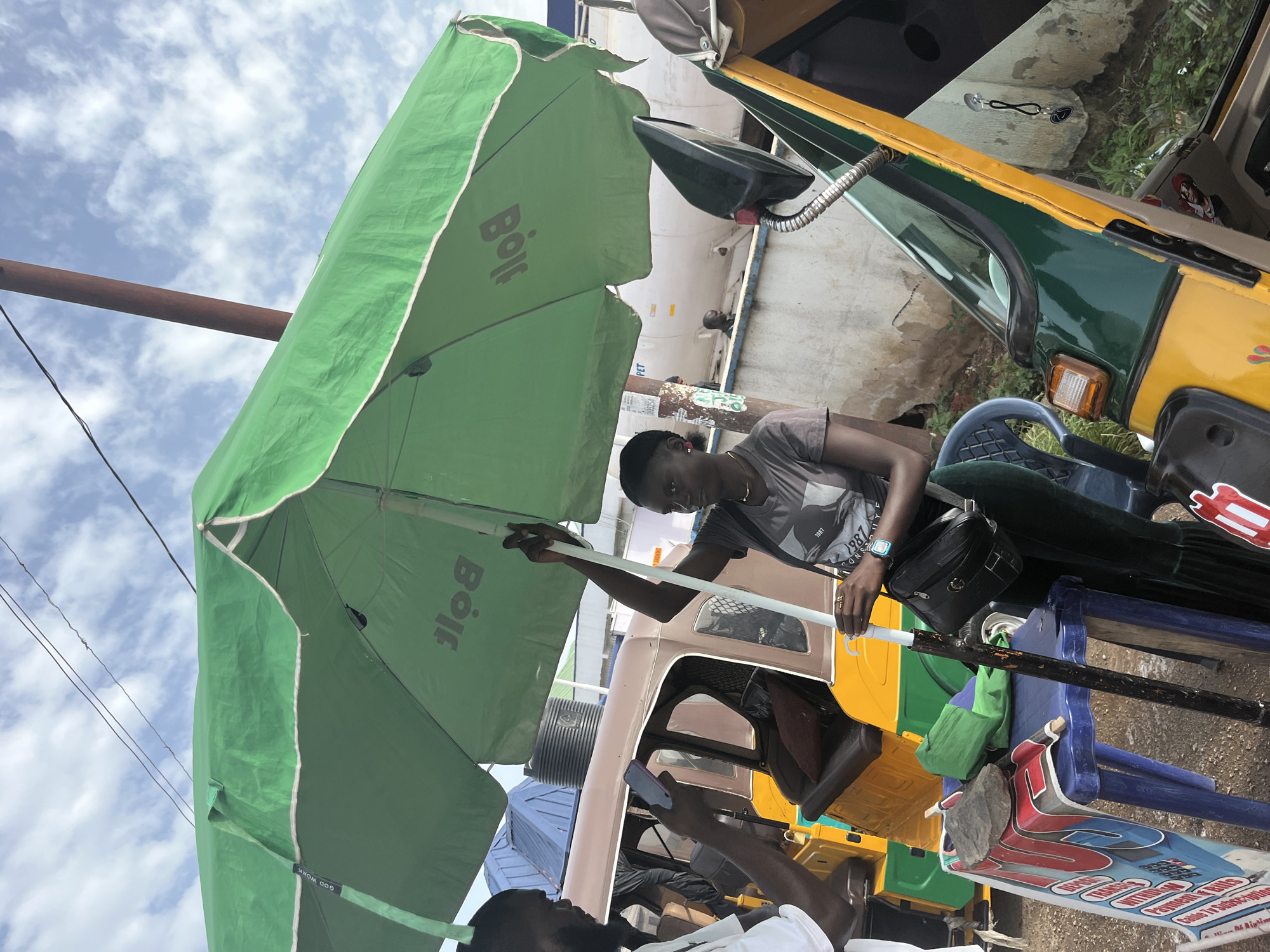 In Jos, Nigeria, a young woman holds an umbrella pole while standing next to a small, three-wheeled vehicle. She wears a grey t-shirt, dark green pants, and a black purse slung across her body. In front of her, a blue plastic table displays a stack of green material. To her left, a man in a white t-shirt holds up a phone. Behind them, another three-wheeled vehicle is parked. The scene is set against a backdrop of a cloudy sky and a large white tank, possibly part of a fuel station. The overall atmosphere suggests a bustling urban environment in Nigeria.