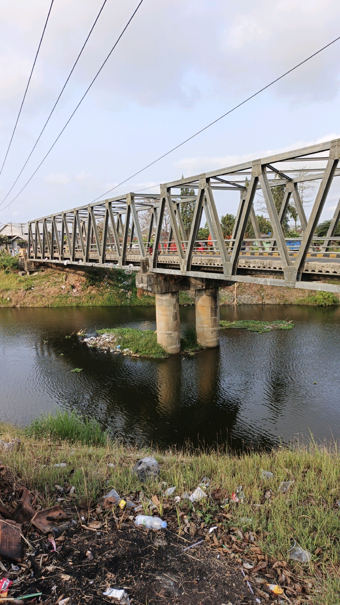 The image depicts a scene in Tanggulwelahan, Indonesia, featuring a truss bridge spanning a body of dark water. The bridge, constructed of grey metal, supports vehicular traffic, with several cars visible on its surface. Two concrete pillars support the bridge from the water. On the right bank of the river, a small island of vegetation is covered in debris, including plastic bottles and other refuse. The riverbanks are lined with grass and scattered litter, suggesting an area with environmental concerns. Overhead, power lines stretch across the frame against a pale sky with scattered clouds. The overall impression is one of an industrial or semi-urban landscape affected by pollution.
