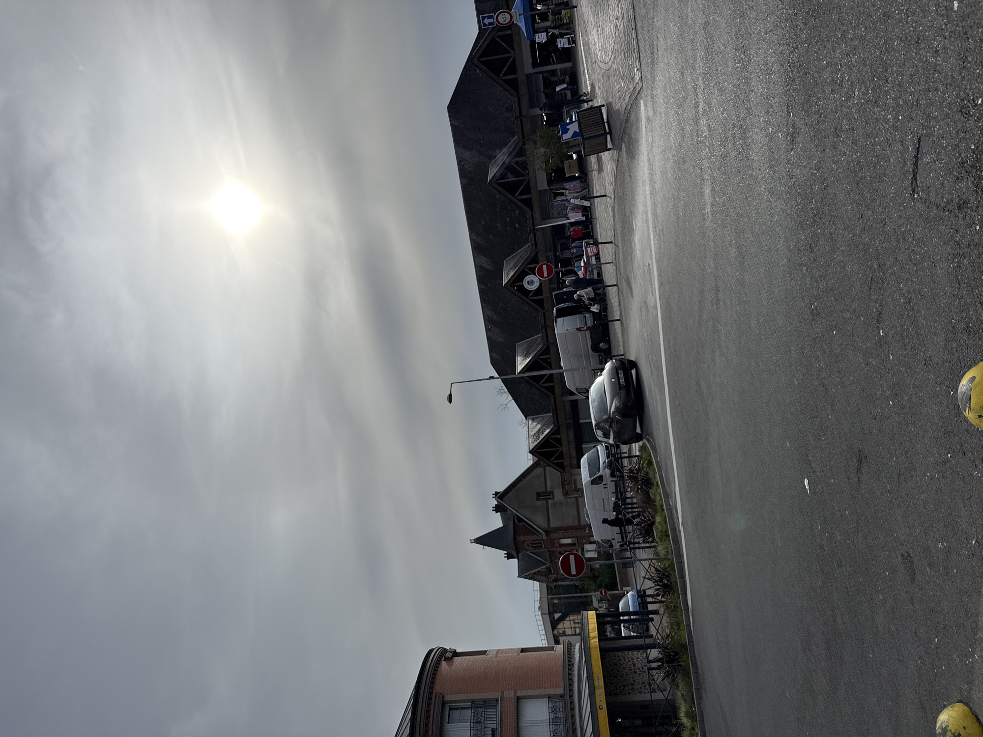 The image depicts a street scene in Domont, France, under an overcast sky with the sun visible through the clouds. The perspective is tilted, showing a downward view of a roadway with a row of buildings along one side. A black asphalt road is in the foreground with a white dividing line. Several parked cars and vans are visible along the curb. One yellow and black parking bollard is in the lower right corner. Buildings on the left display various architectural styles, including gable roofs. Traffic signs indicating restrictions are present. A lamppost is also visible. There are no visible people or immediate activity apparent in the frame.