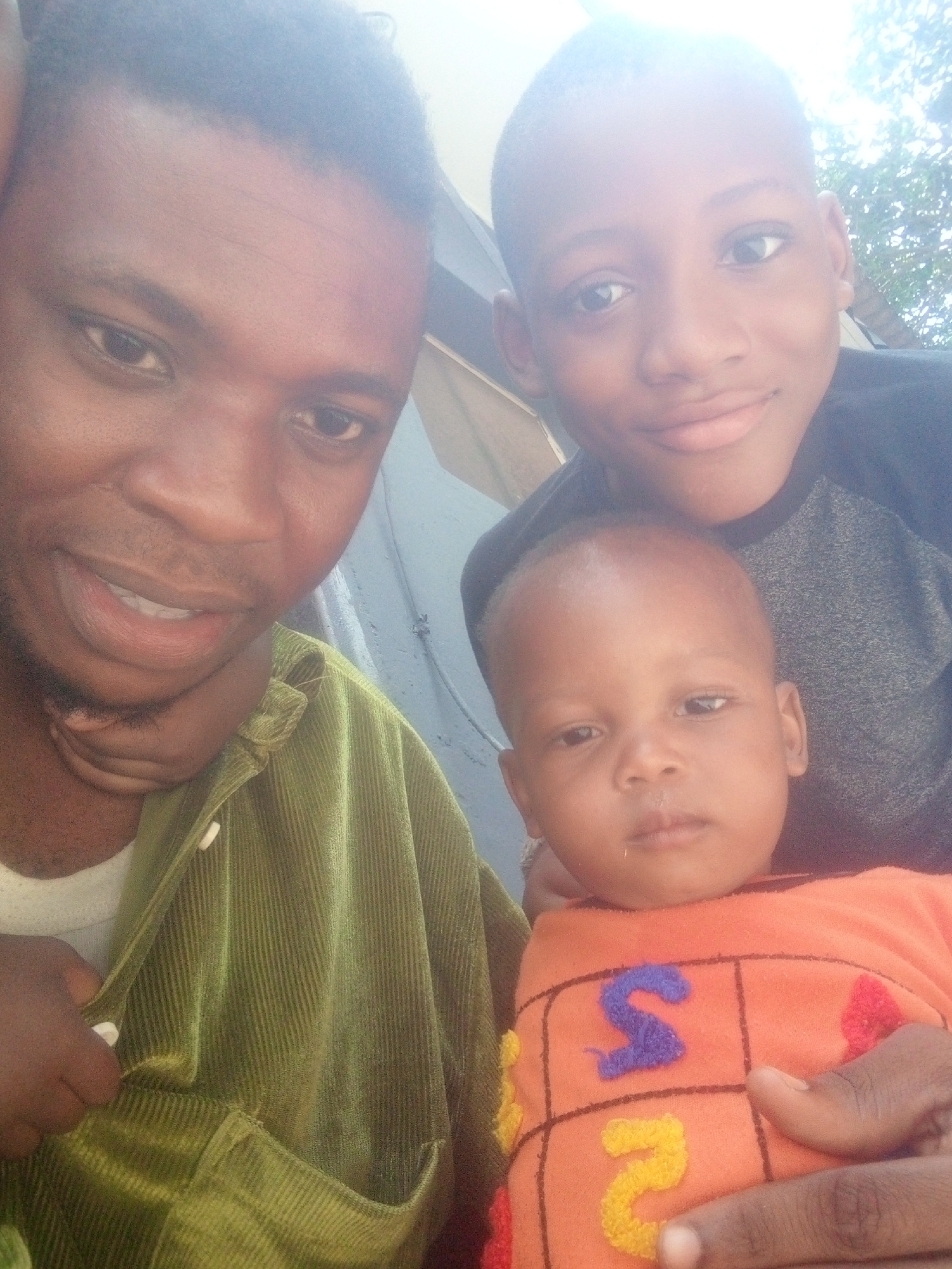 A bright, close-up selfie-style photograph captures three males of varying ages, likely a family, posing together. The subjects are all of African descent. On the left, a man with dark skin, short dark hair, and light stubble looks towards the camera with a slight smile. He is wearing an olive green, corduroy-textured jacket over a white undershirt, with white buttons visible on the jacket. His left hand is resting under his chin.
To the right and slightly behind him is an older boy, also with dark skin and short dark hair, smiling broadly at the camera. He is wearing a black and grey two-tone t-shirt. Below him, in the foreground and center, is a baby or toddler with dark skin and very short hair or bald, looking directly at the camera with a neutral expression. The baby is wearing a vibrant orange long-sleeved top that features colorful, embroidered letters and shapes. Clearly visible are a purple "S" and a yellow "2", along with parts of red embroidered elements.
The lighting is very bright, almost overexposed in the upper right portion of the image, suggesting an outdoor setting in strong daylight. Hints of foliage or trees are visible in the bright background, along with what appears to be a light-colored wall or structure. The overall impression is one of a warm, familial moment.