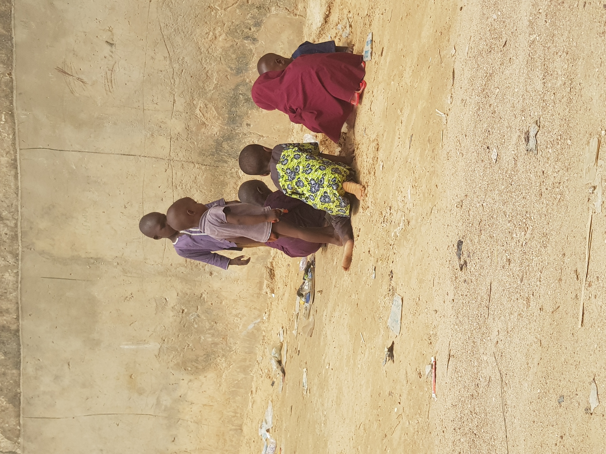 In Dutsinma B, Nigeria, a group of children gather on a sandy patch of ground. A wall runs along the left side of the frame. The children are dressed in various colorful clothes, and some are barefoot. Debris litters the sand.