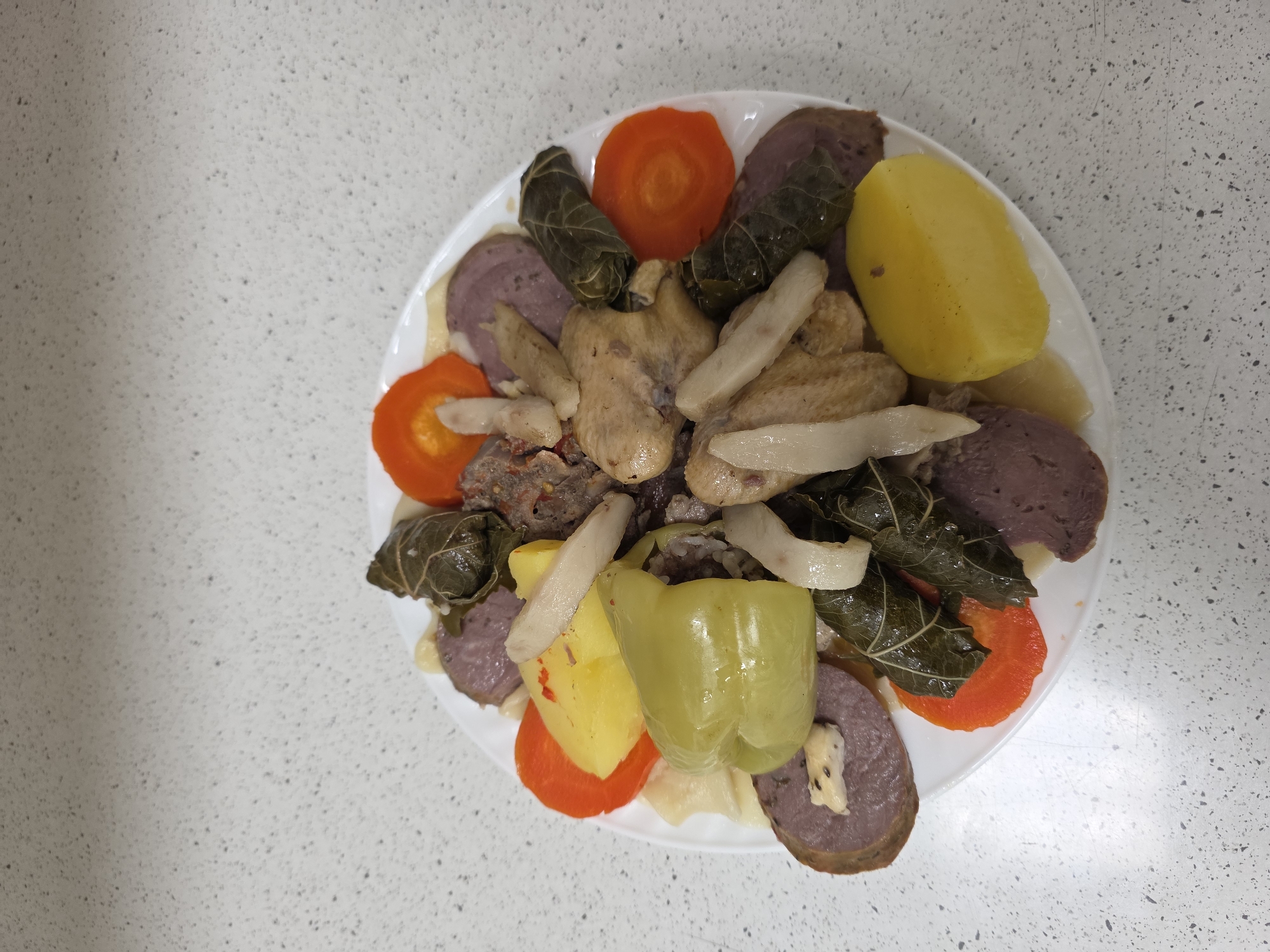 A white plate of food rests on a speckled white countertop. The plate is filled with a variety of food items including sliced carrots, what appear to be dolmades or stuffed grape leaves, pieces of cooked chicken, sliced purple sausage, boiled potatoes, and a large green bell pepper. The food is arranged in a somewhat haphazard manner. The background is a close-up of a countertop with small black speckles. The image is taken from a top-down perspective. There are no people or identifiable city locations visible in the image.